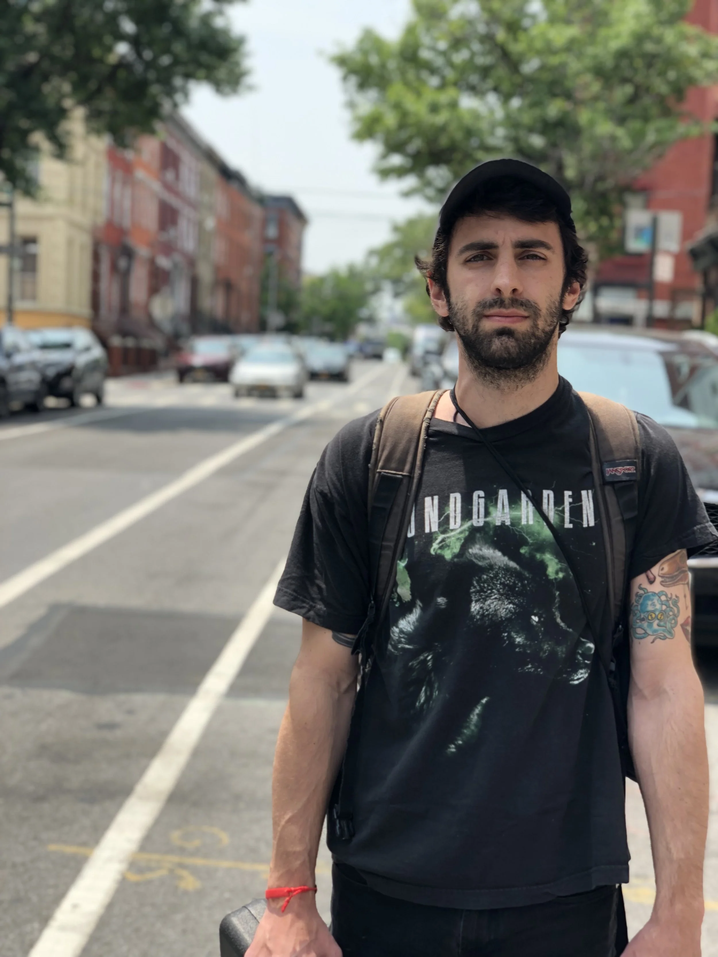 A man with dark hair and a beard wearing a black Nirvana T-shirt and a black cap, standing on a city street with parked cars, trees, and brick buildings in the background.