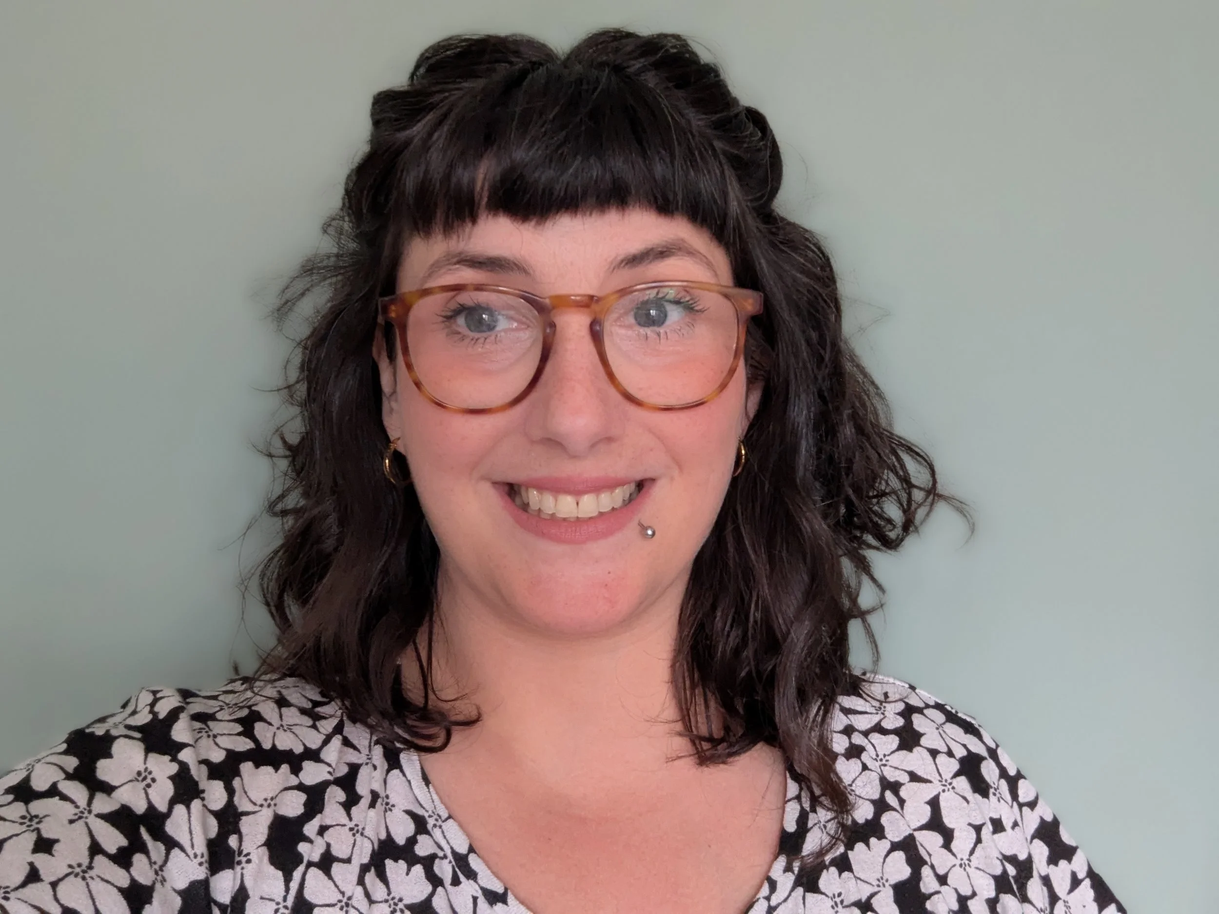 Smiling woman with dark hair, bangs, orange glasses, lip piercing, and a black-and-white floral top