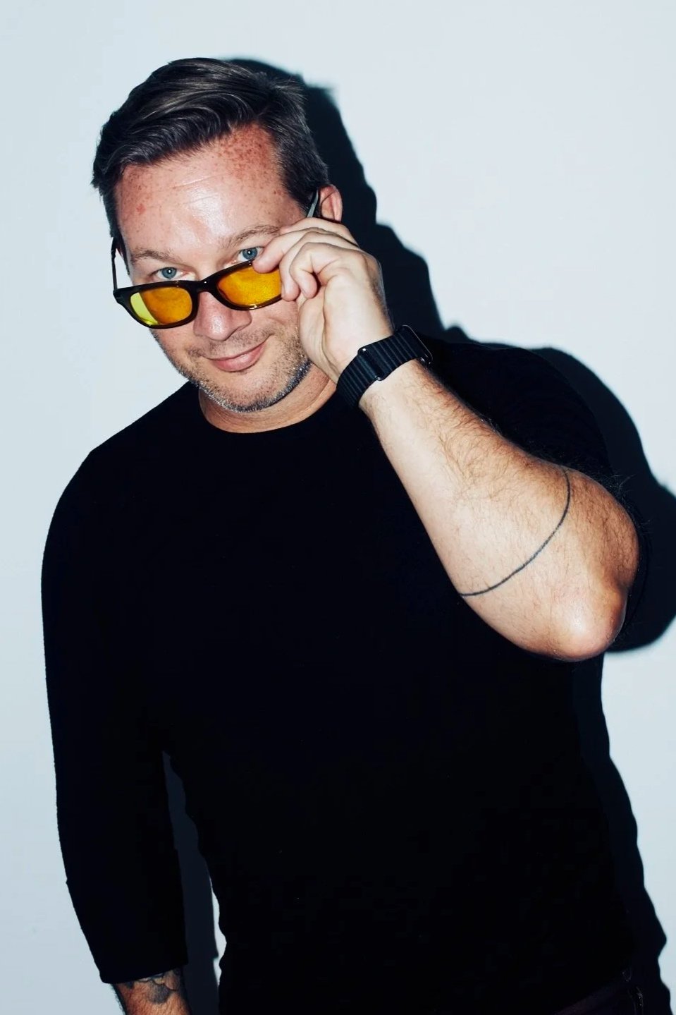 Man in a black shirt adjusting amber-tinted sunglasses against a light wall