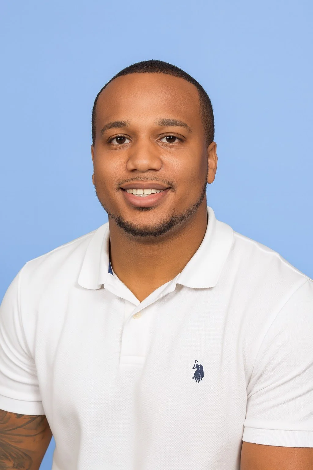 A young man with short hair, a beard, and a smile, wearing a white polo shirt against a light blue background.