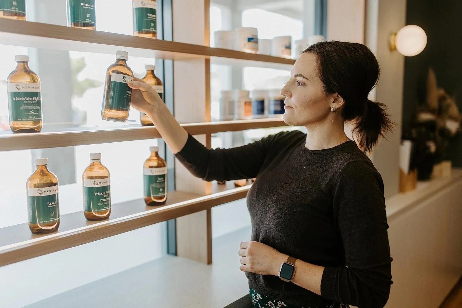 A woman in a black long-sleeve shirt selecting a bottle from a wooden display shelf with other labeled bottles.