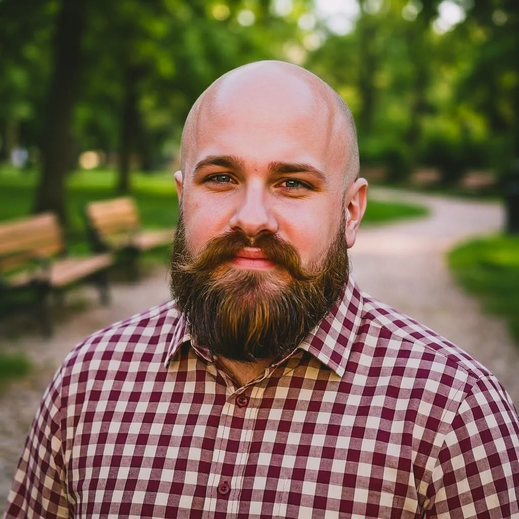 A man with a beard and mustache, wearing a red and white checkered shirt, standing outdoors in a park with green trees and benches in the background.