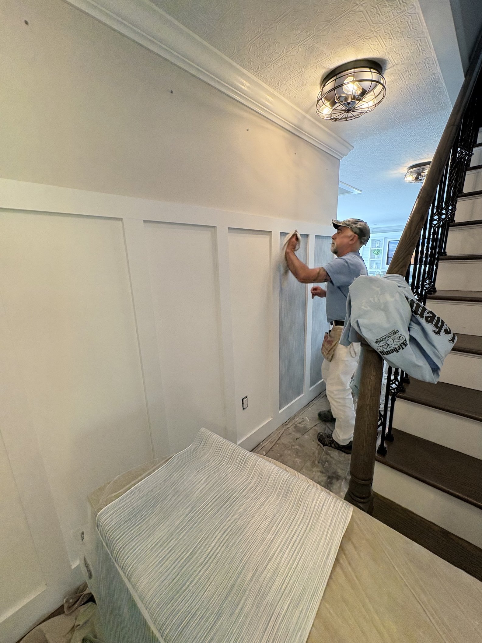 A man in a gray shirt and khaki pants is sanding a white wainscoting panel on the wall near a staircase. There is a striped upholstered bench in the foreground.