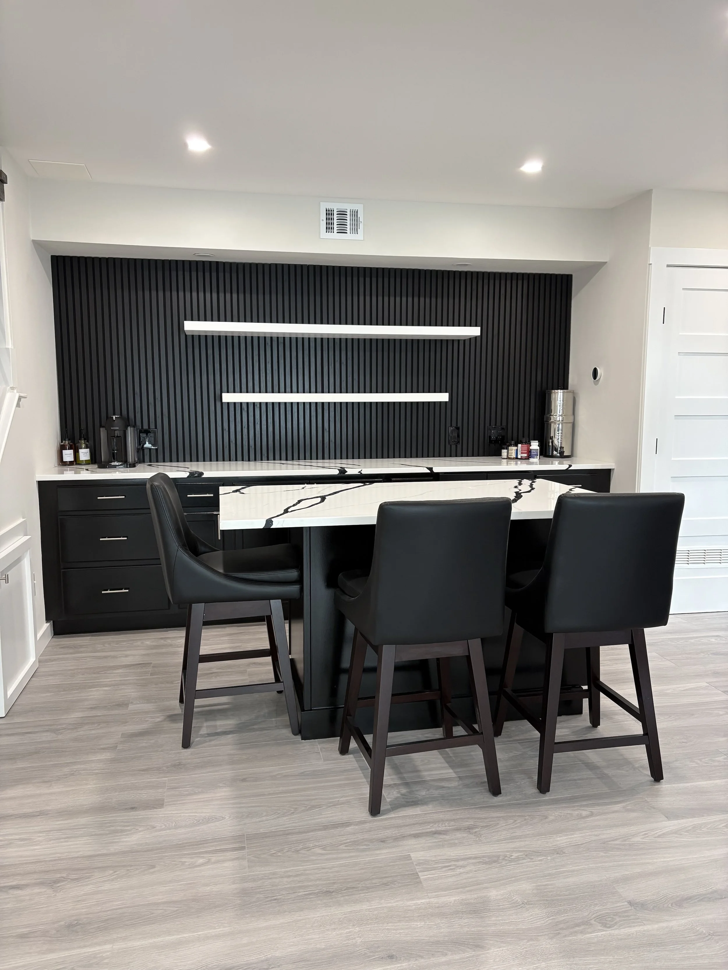 Kitchen with black and white island, black chairs, black textured wall with two white shelves, and small appliances and decor on the countertop.