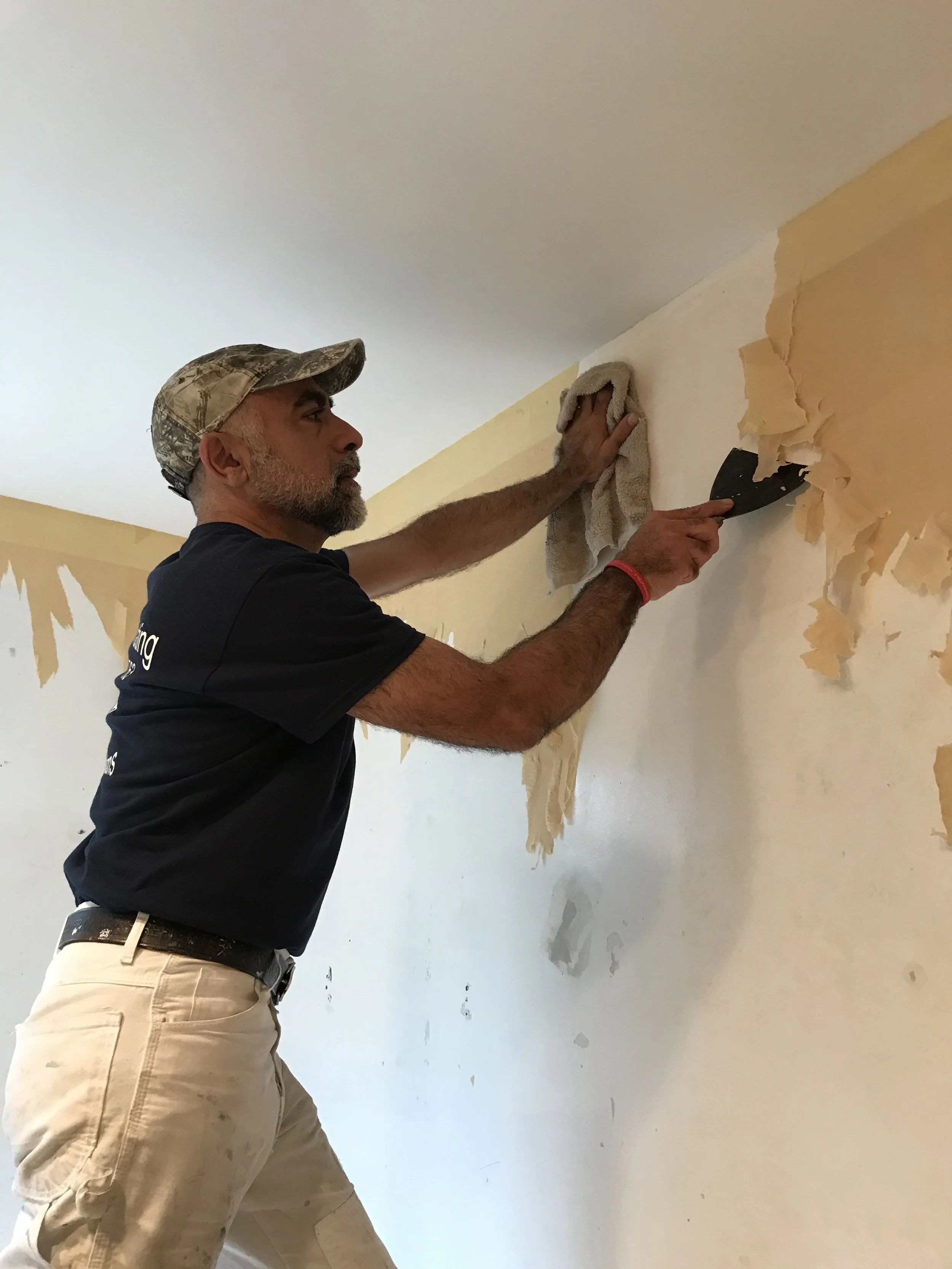 A man wearing a camouflage cap and khaki pants is using a putty knife to scrape wallpaper off a wall in a room with some damaged paint and wallpaper in Baryville New York
