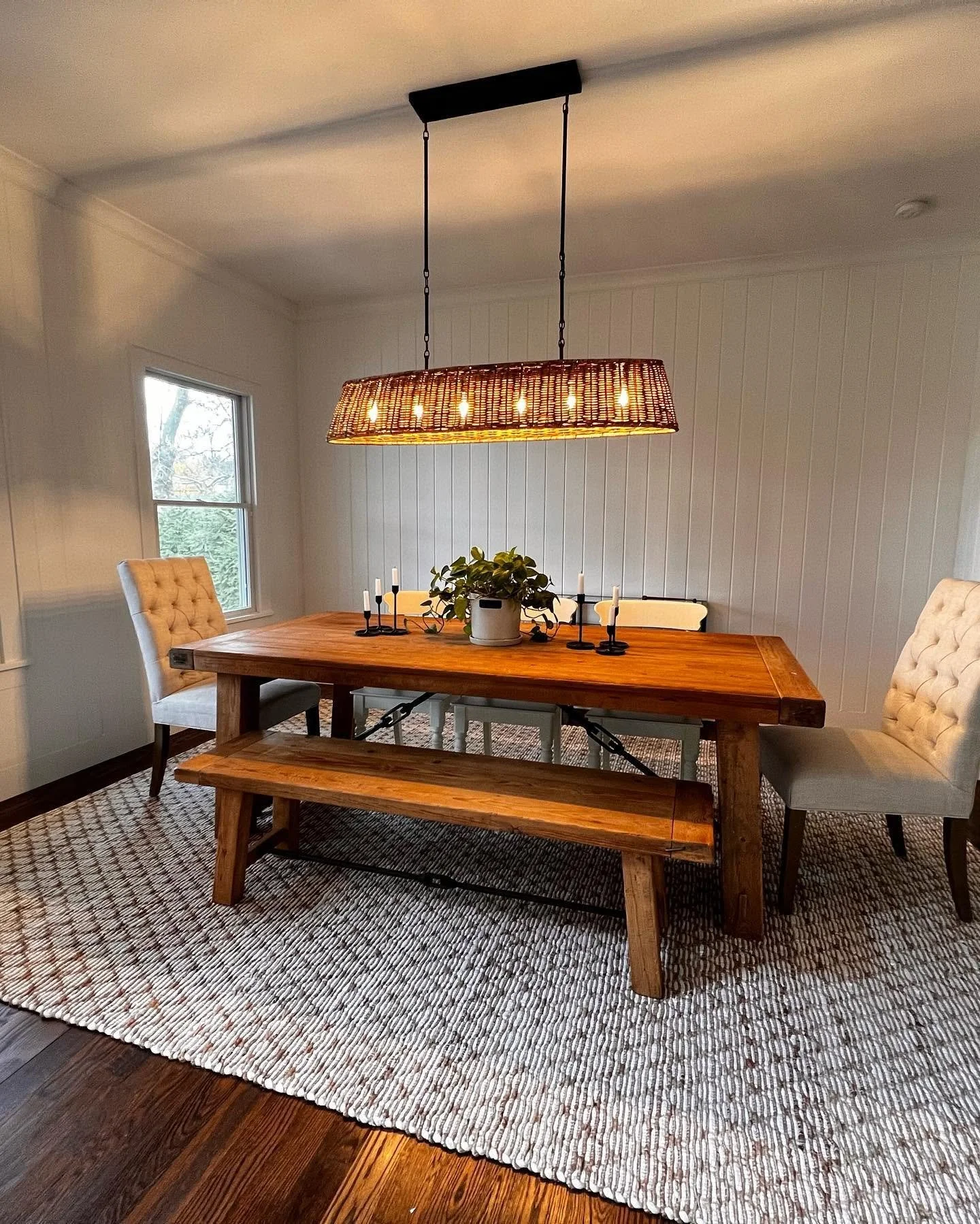 A dining room with a wooden table and matching benches, surrounded by white upholstered chairs. A large woven light fixture hangs above the table, and a potted plant with white candles is in the center. The room has a window on the left with a view of trees outside, white beadboard walls, and a textured rug on dark wooden floors.