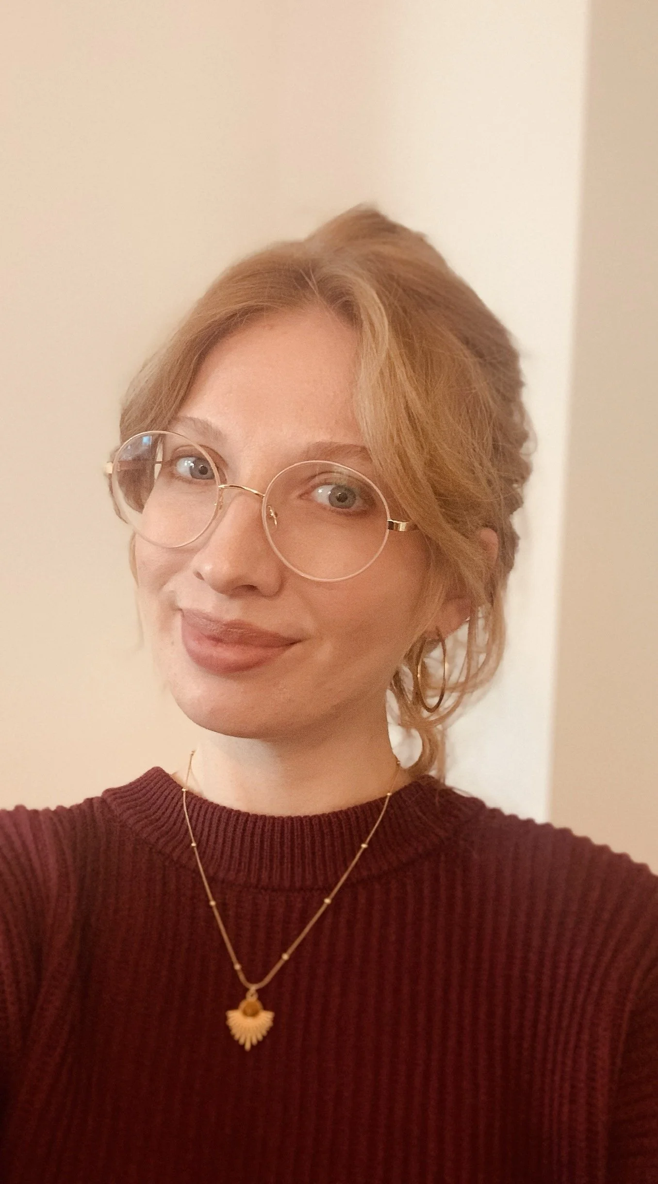 Porträt einer jungen Frau mit rötlich-blonden, lockigen, kurz geschnittenen Haaren, rundlichen Brillengläsern, einem dunkelroten Pullover, goldener Kette mit Anhänger und dezentem Makeup, lächelnd vor neutralem Hintergrund.