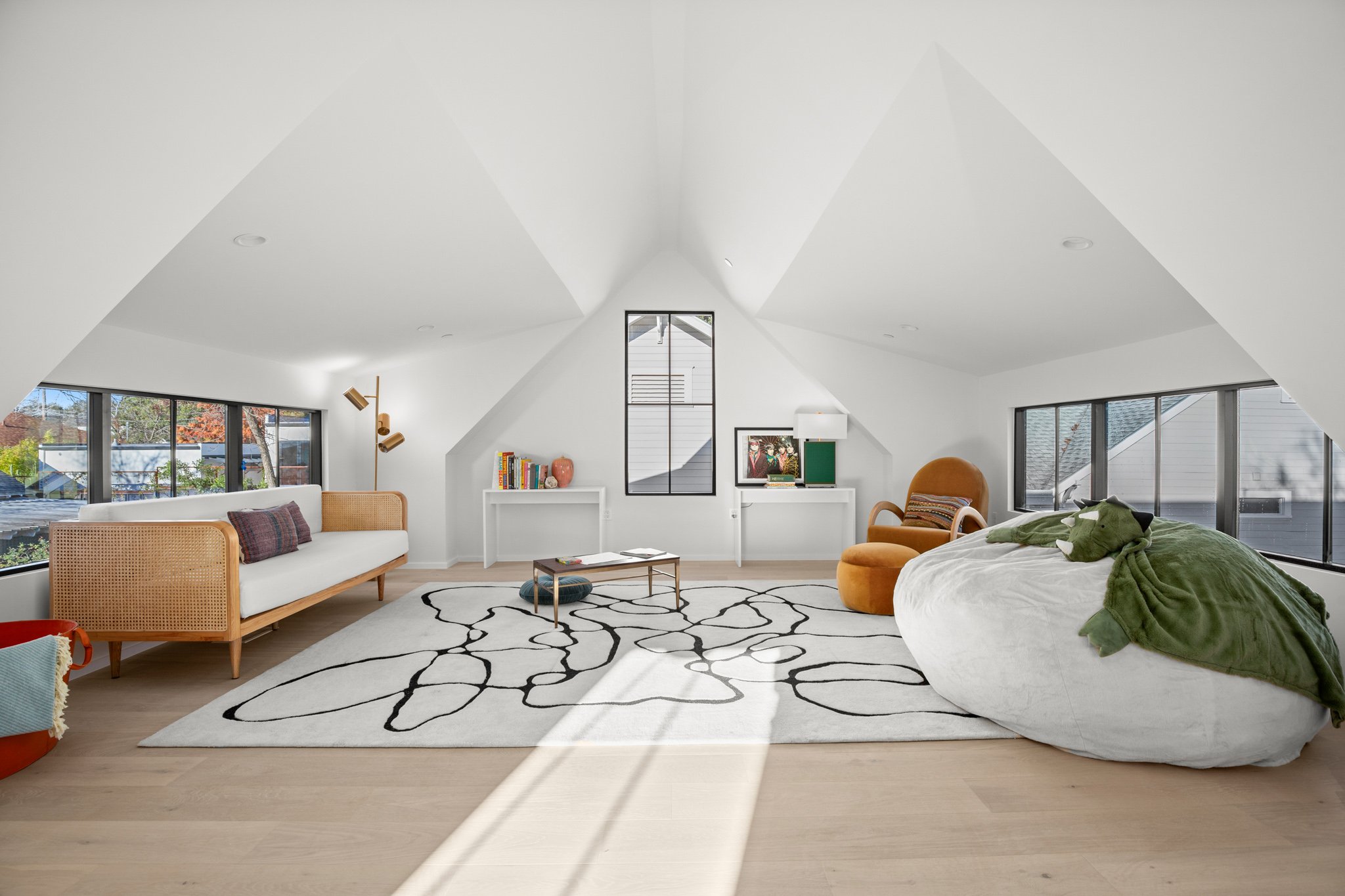 Spacious modern bonus room with white vaulted ceilings, large windows, a white sofa with plaid pillows, a patterned area rug, a round green chair, a bean bag with green blanket, and artwork on the wall. The Kids' escape room.