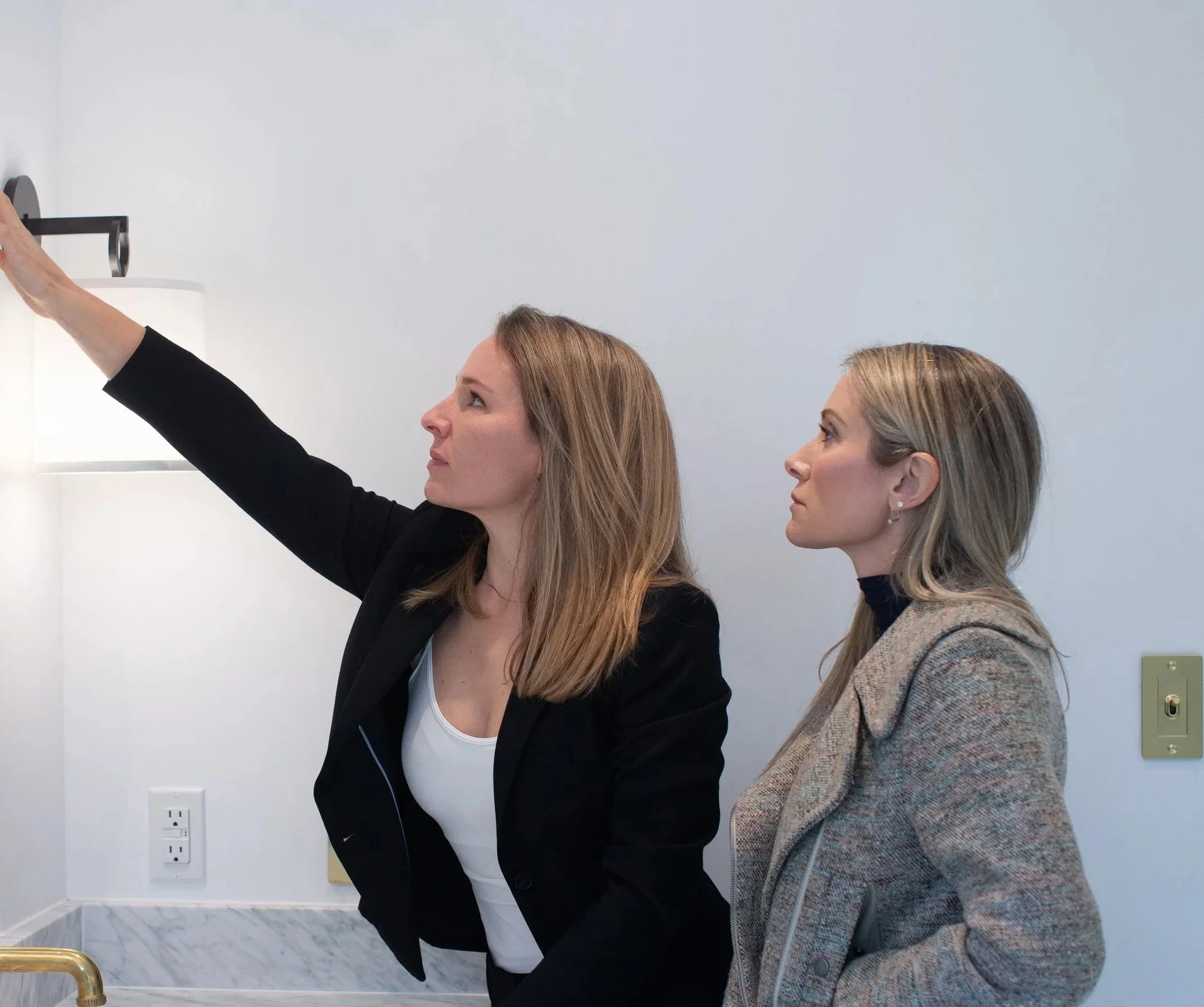 Two design consultants observing a wall-mounted lamp in a modern room with white walls and marble flooring.