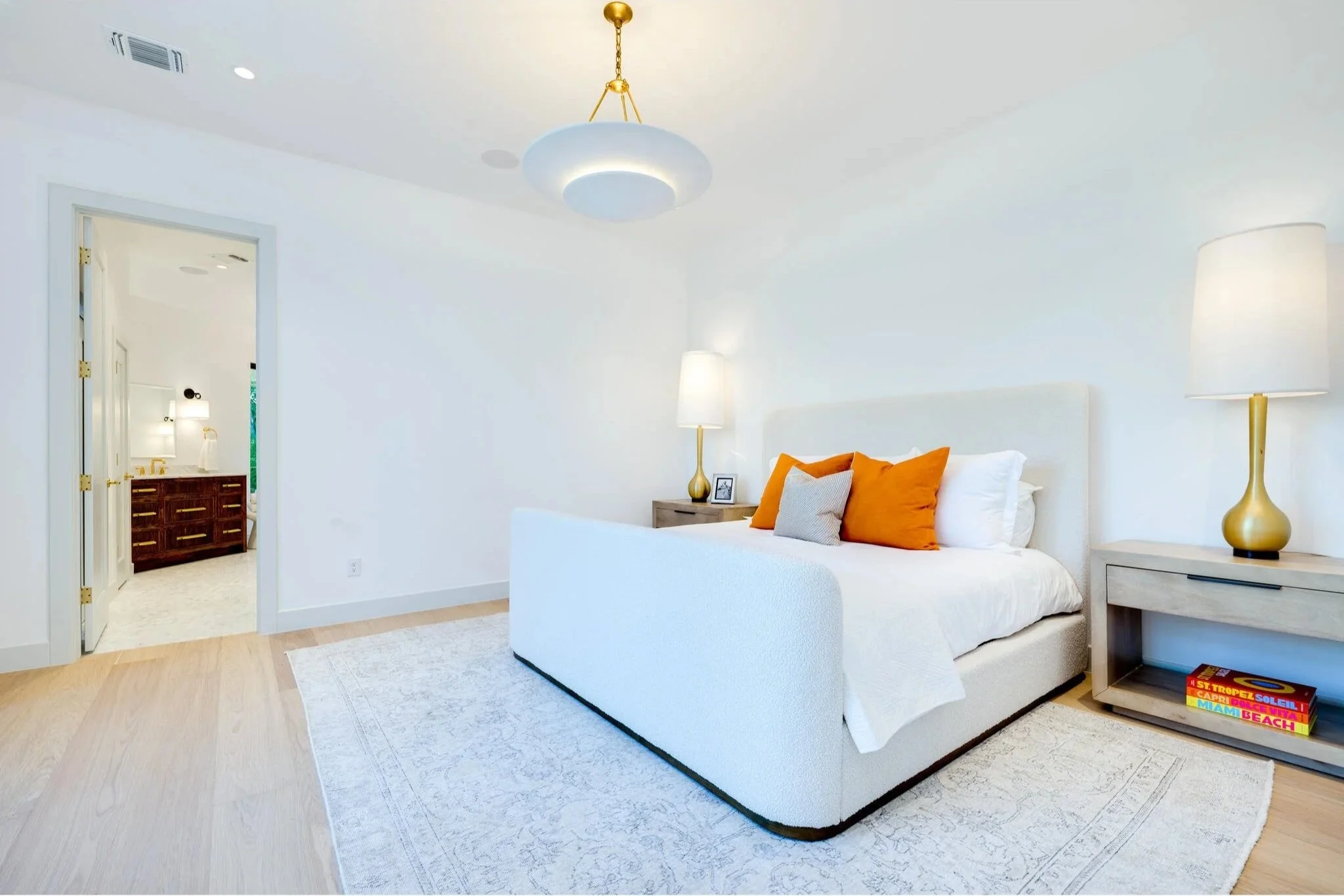 A bright, white bedroom with a large bed adorned with orange, white, and gray pillows, illuminated by two tall matching lamps on either side. A modern hanging light fixture with a gold chain is suspended from the ceiling. On the right side, a bedside table with a colorful stack of books is visible. The bedroom has light wood flooring and a textured white rug. A doorway leads to a bathroom with dark wood cabinetry and white vanity.