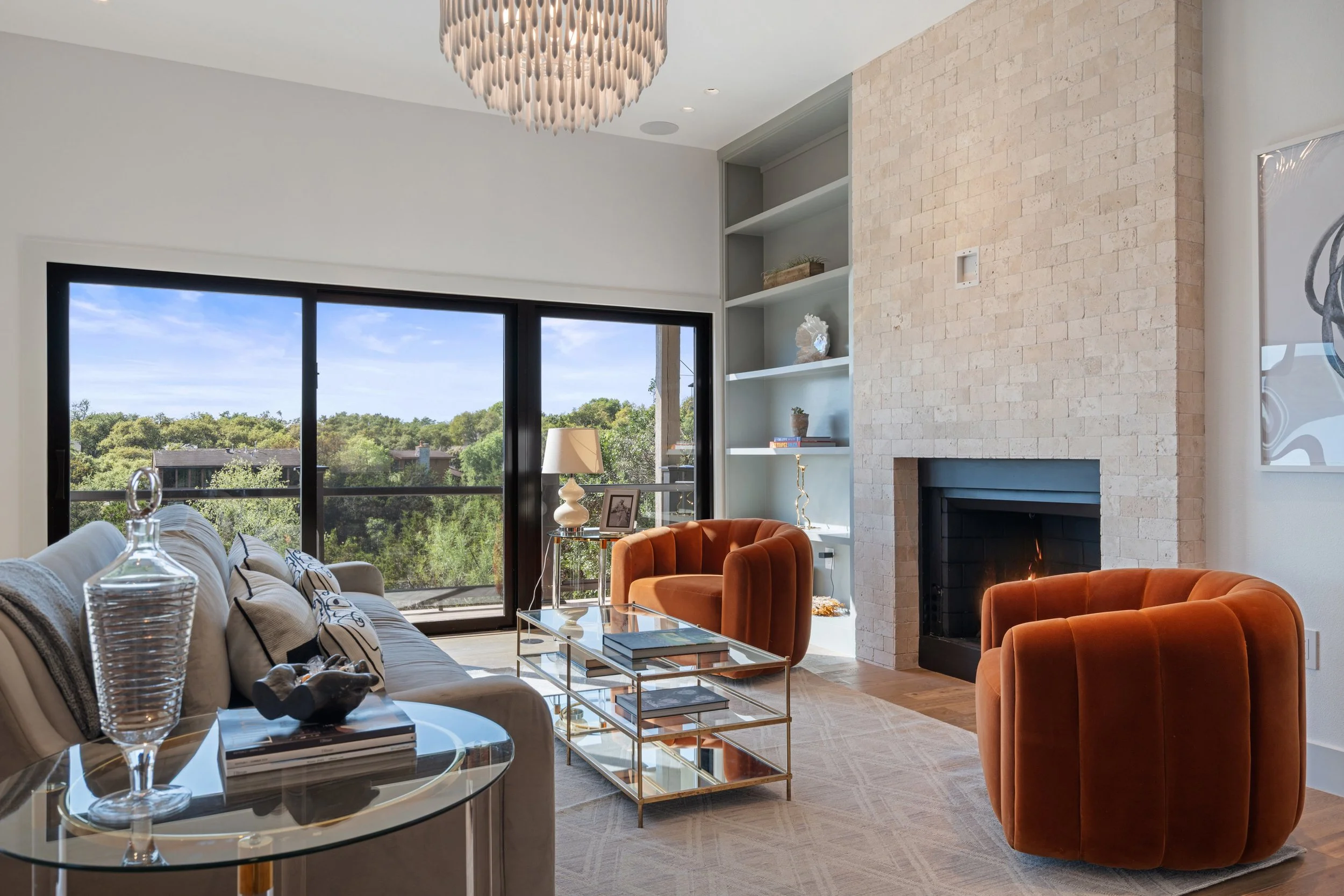 Living room with sliding glass doors showing the hill country, curated furniture showcases a soft grey sofa with pillows, brass glass coffee table, orange velvet chairs, limestone fireplace, built-in shelves with decor, and visual comfort lighting.