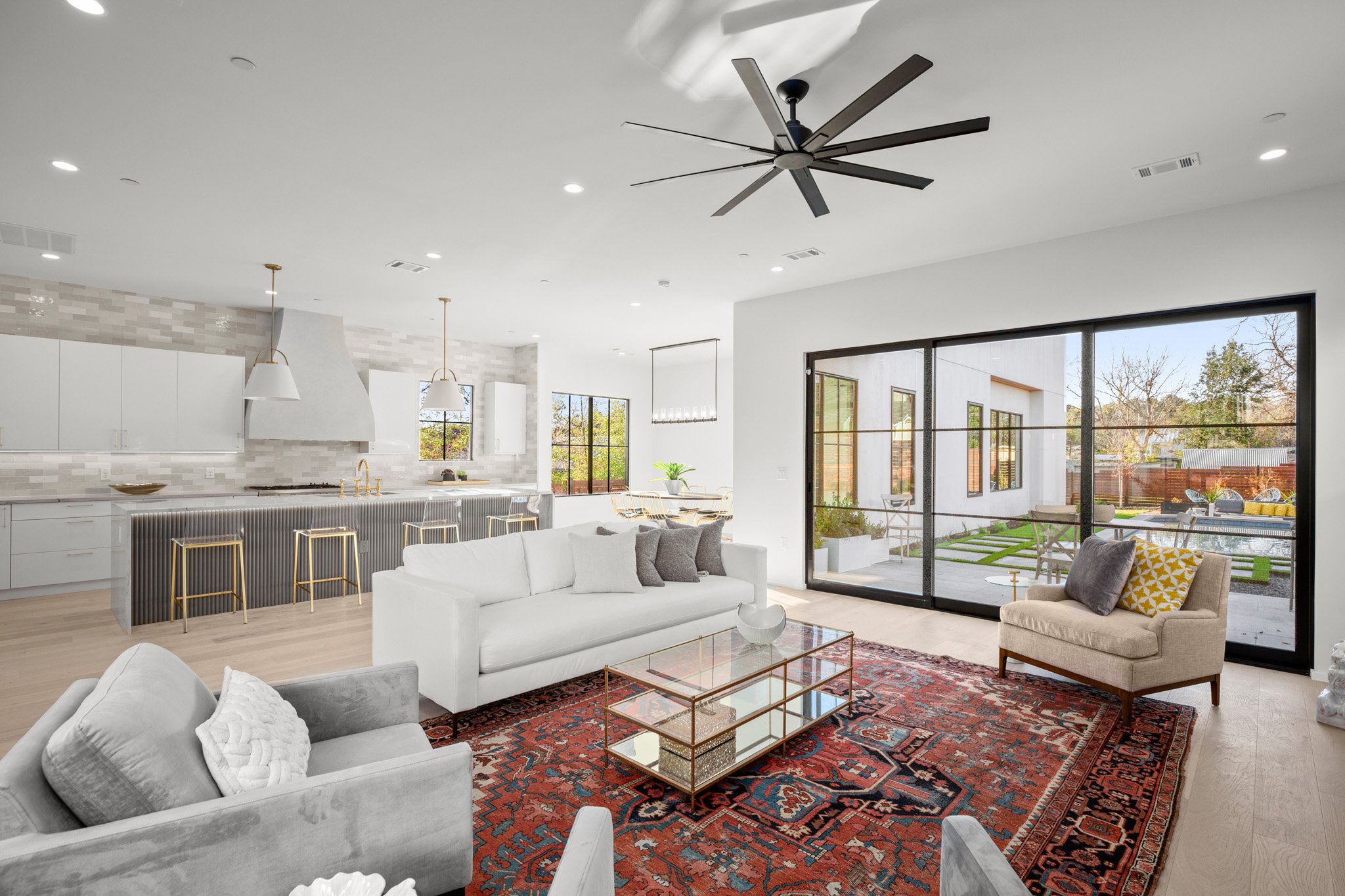 Open-concept living area with white sofas, a decorative oriental rug, glass coffee table, and large sliding glass doors opening to a backyard patio with seating and a grassy area.