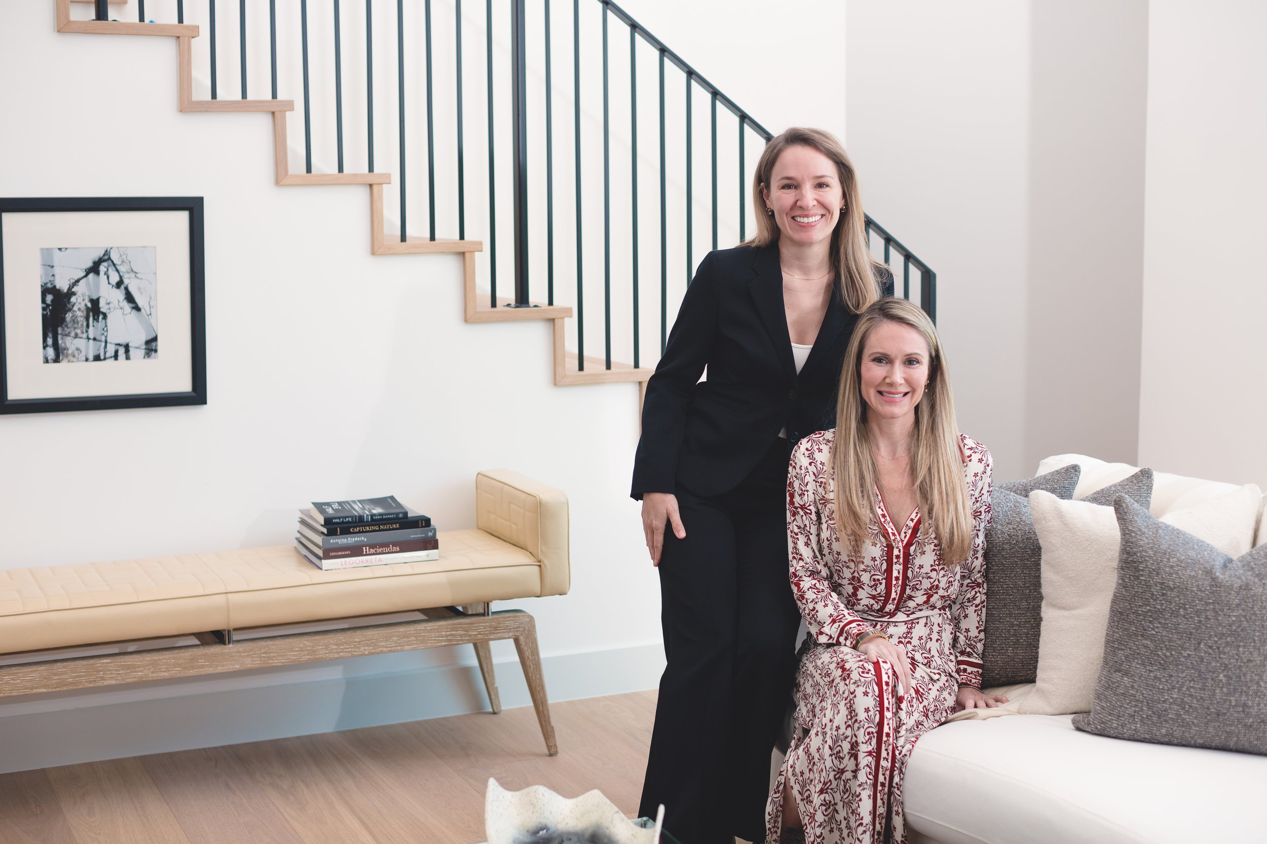 Two women in a design focused living room. The room has white walls, black railing stairs, a framed black and white photo on the wall, a small stack of books, a cream bench, and pillows on the sofa.
