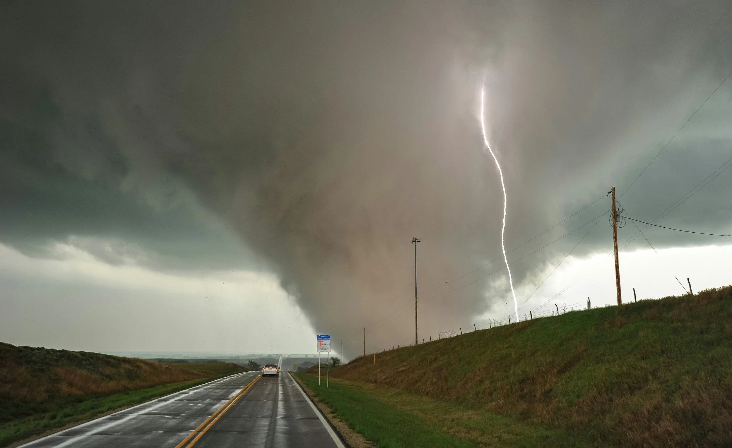 A lightning bolt striking through a cloudy sky over a rural road with a car driving away, utility poles lining the side of the road, and a signpost in the distance.