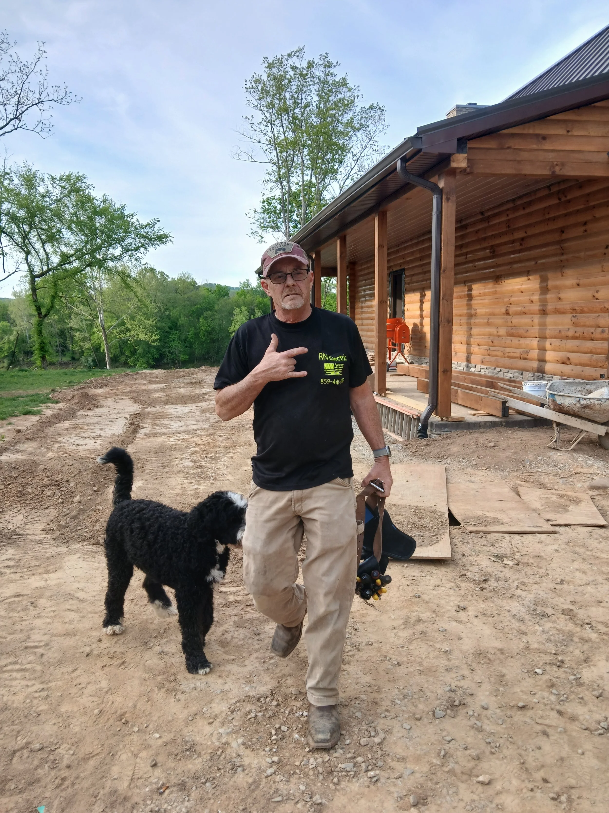 A man wearing glasses, a pink cap, black T-shirt, and khaki pants walking on a dirt path next to a black and white dog. The man is making a gesture with his hand pointing to his chest. Behind him is a wooden house under construction with trees in the background.