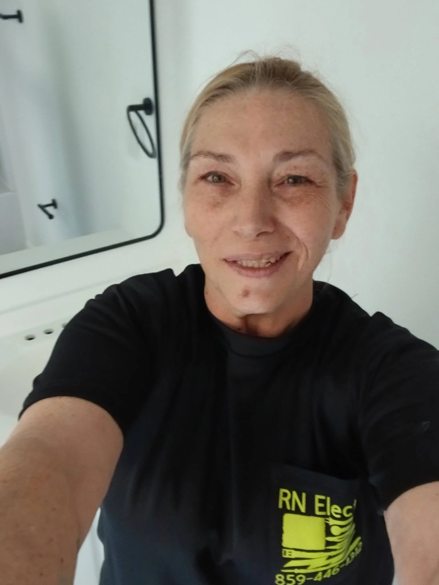 A woman taking a selfie in a mirror, wearing a black t-shirt with yellow text. The woman has a big smile and her hair is pulled back. The background includes a mirror, a white wall, and part of a white medical or safety cabinet.