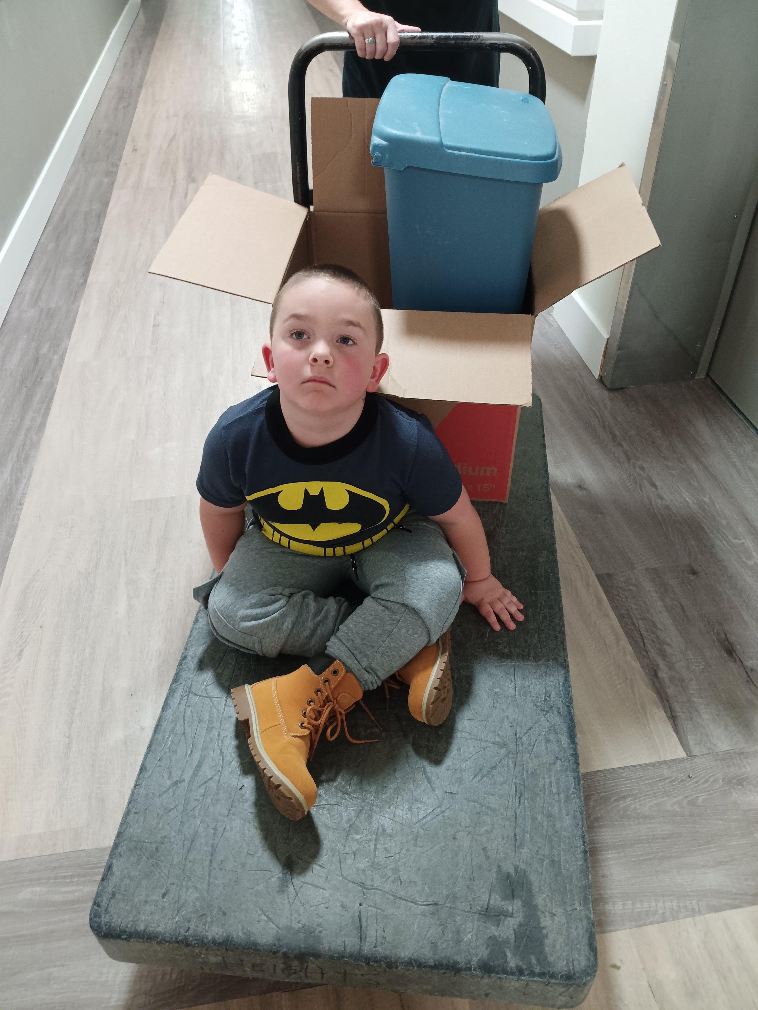 A young boy is sitting cross-legged on a bench with his hands on his lap. He is wearing a Batman T-shirt, gray sweatpants, and tan Timberland boots. There is a large open cardboard box behind him with a blue recycling bin inside.