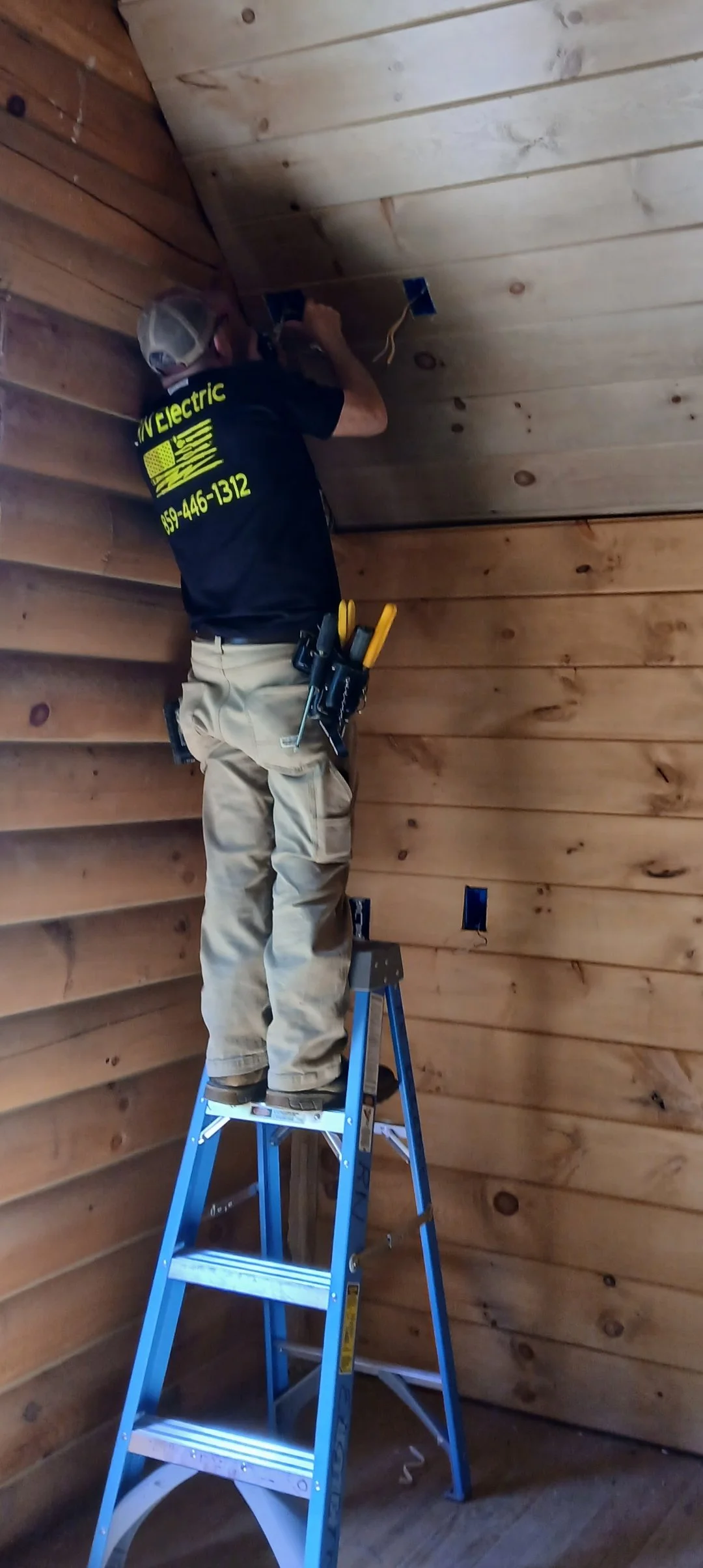 A worker standing on a blue stepladder installing an electrical outlet in a wooden-paneled room; the worker is wearing a black shirt with yellow text, khaki pants, and a utility belt.