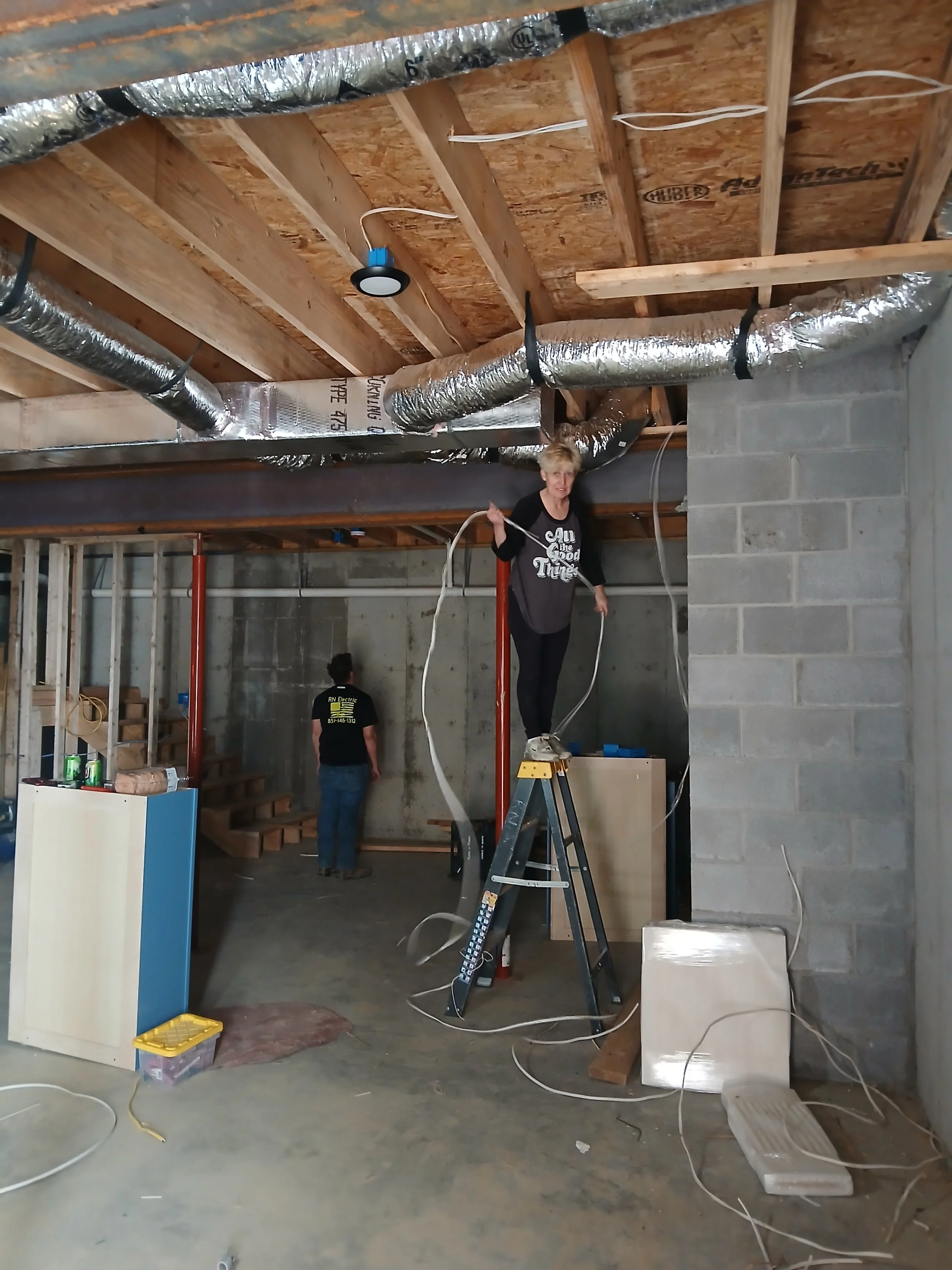 Construction site with exposed ceiling beams, ductwork, and electrical wiring, featuring a woman standing on a ladder, holding a wire, and two other workers in the background.