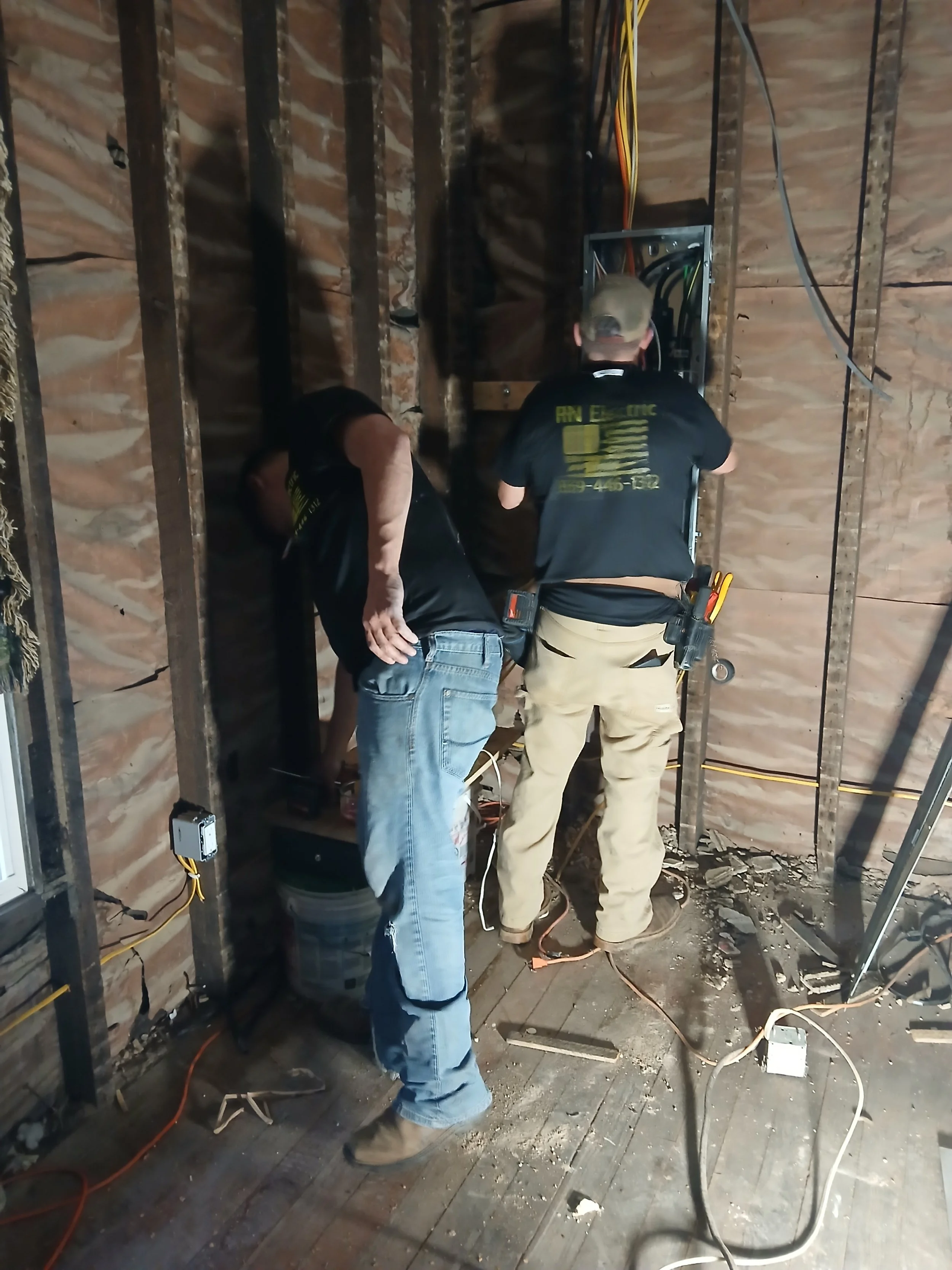 Two workers are installing or repairing electrical wiring inside a building with exposed wooden wall studs. One worker is wearing a black shirt with yellow text and the other is wearing a tactical belt with tools.