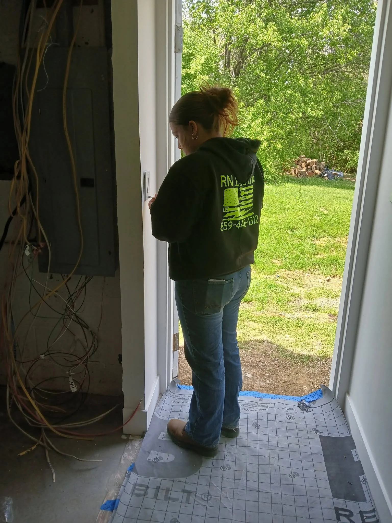 A person standing near a doorway and looking at a device, with construction materials and wiring inside a building and a view of green trees outside.