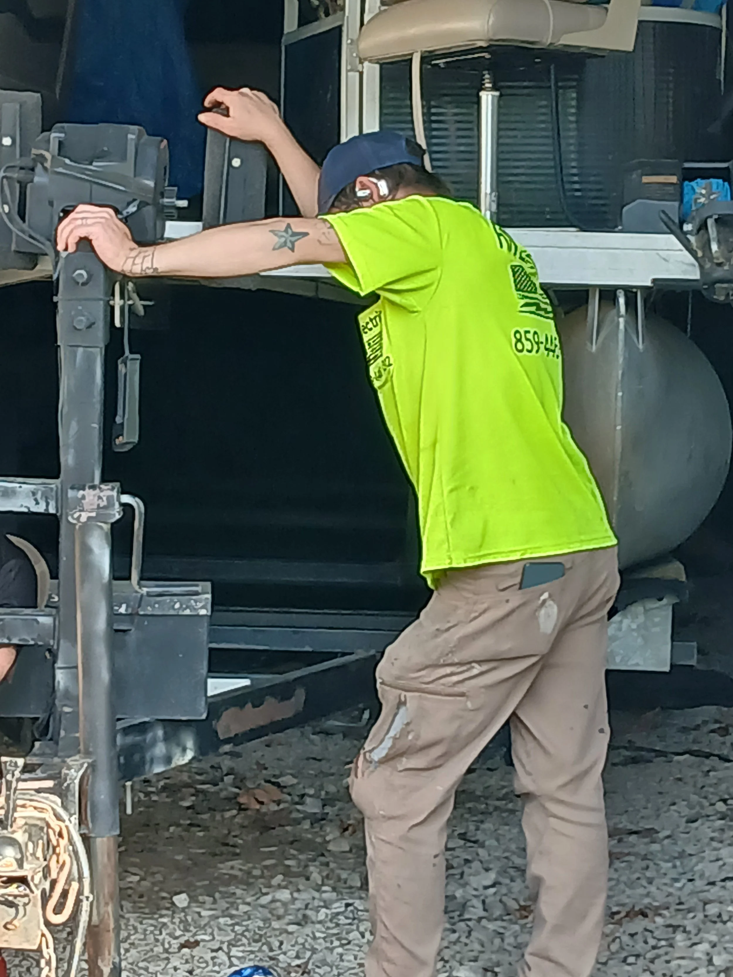 Worker in a yellow safety vest and beige pants working on construction or maintenance at a site with gravel ground, machinery, and equipment.