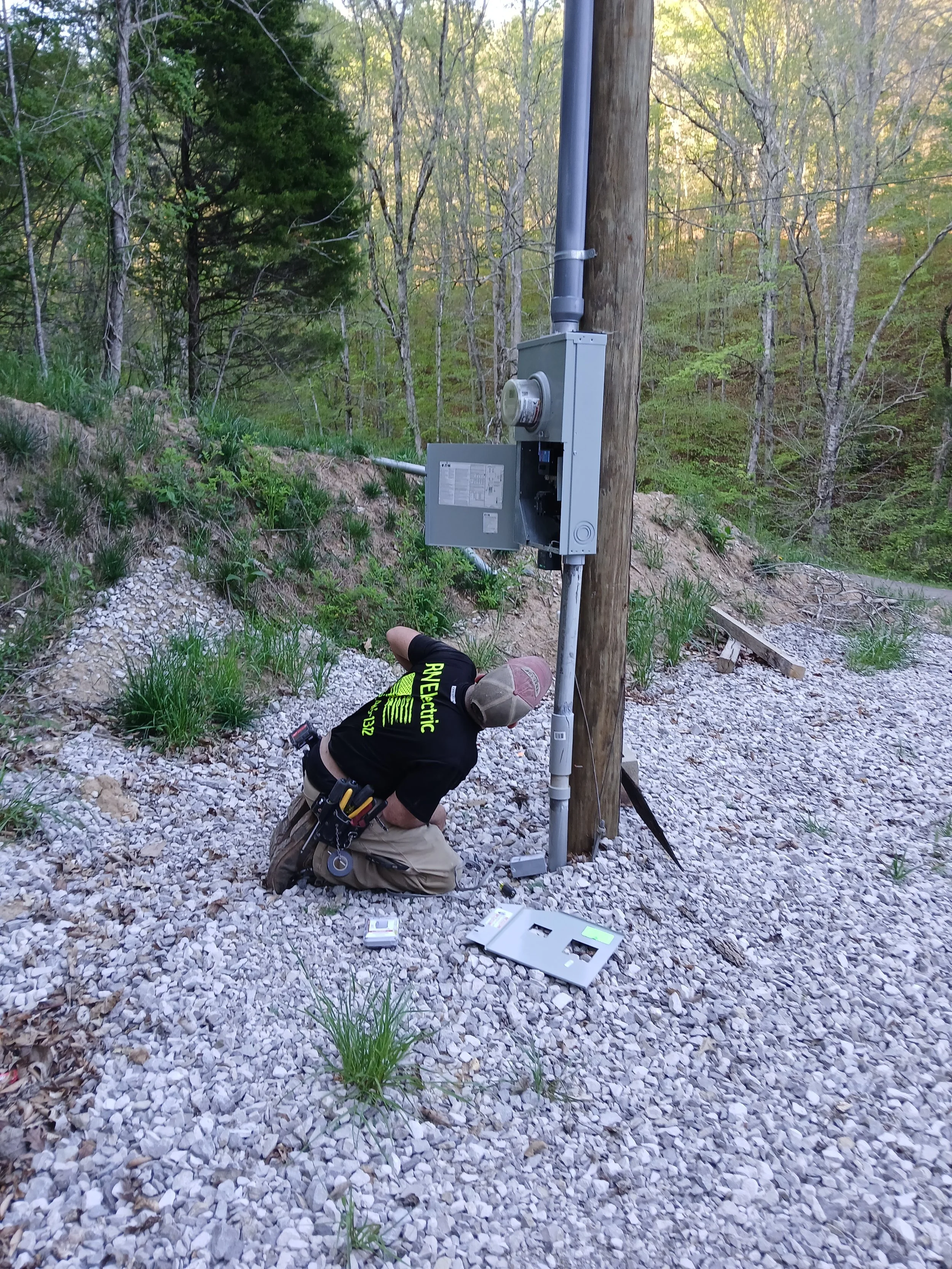 A worker kneeling on a gravel path installing or repairing electrical equipment on a utility pole in a forested area, with tools and electrical panels on the ground.