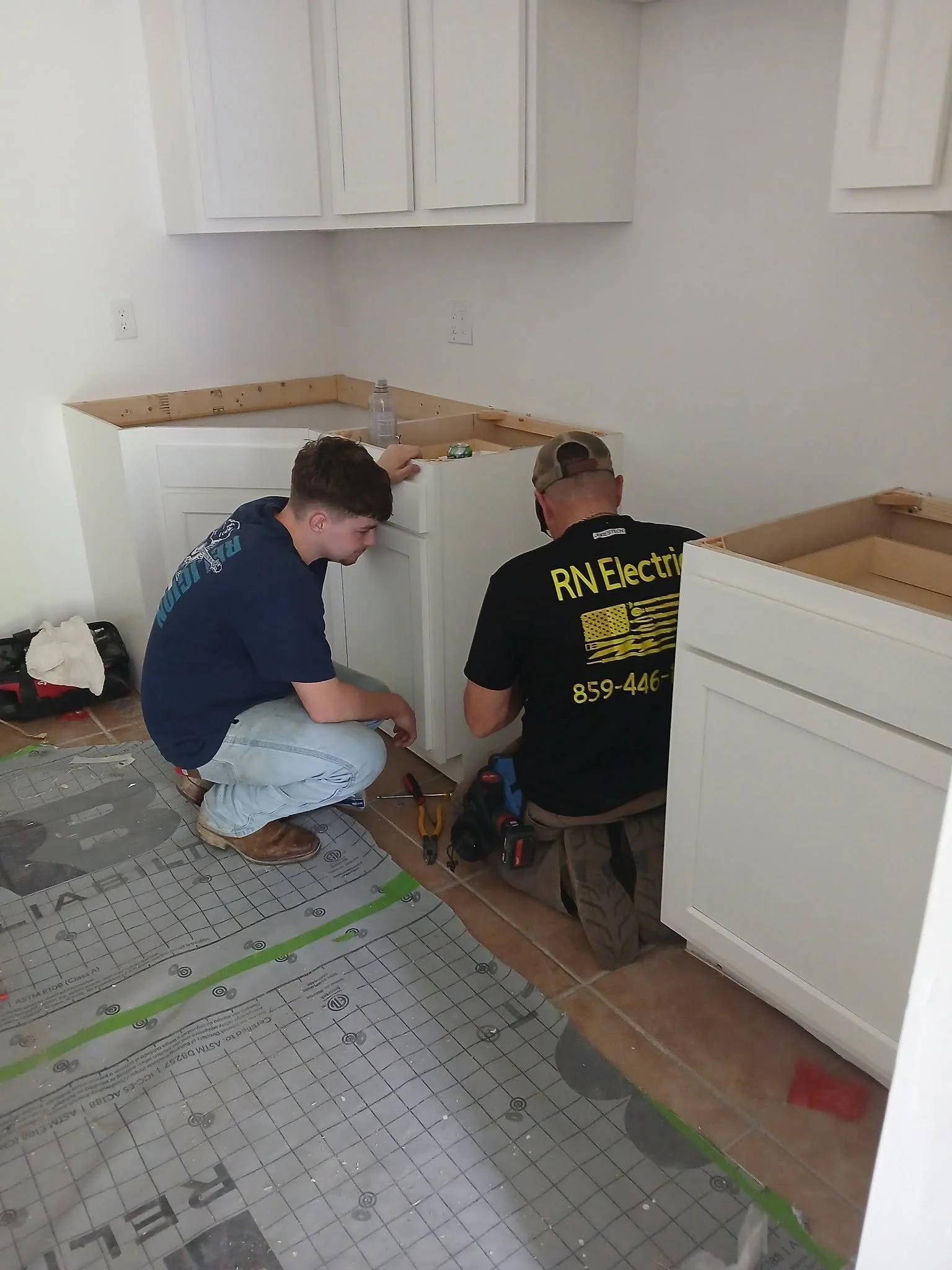 Two people working on kitchen cabinet installation, with tools on the floor and a sheet of underlayment on the floor.