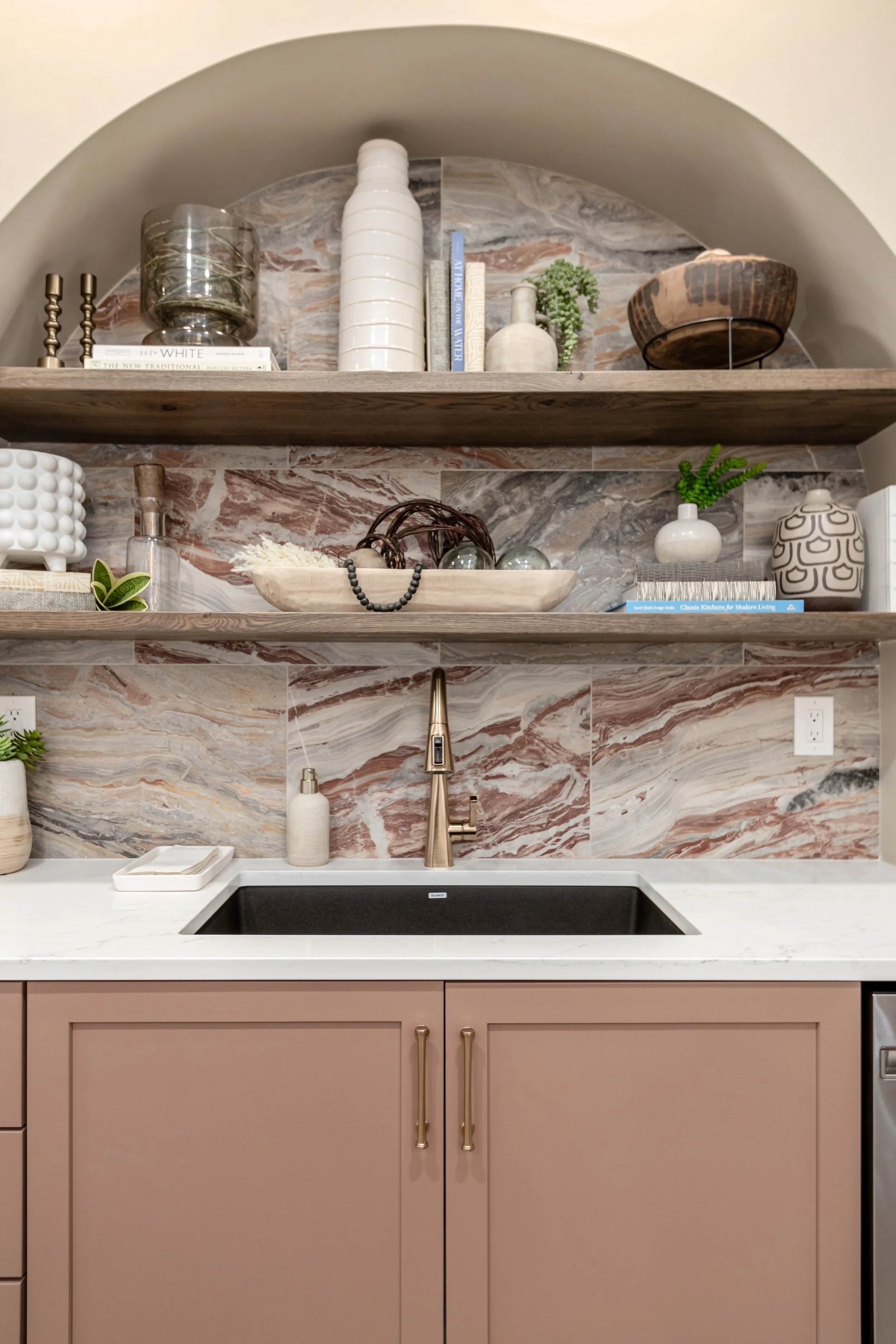 Close-up of a kitchen countertop with a sink, beige cabinet doors, decorative open shelves with vases, books, and decorative items, against a marble backsplash.