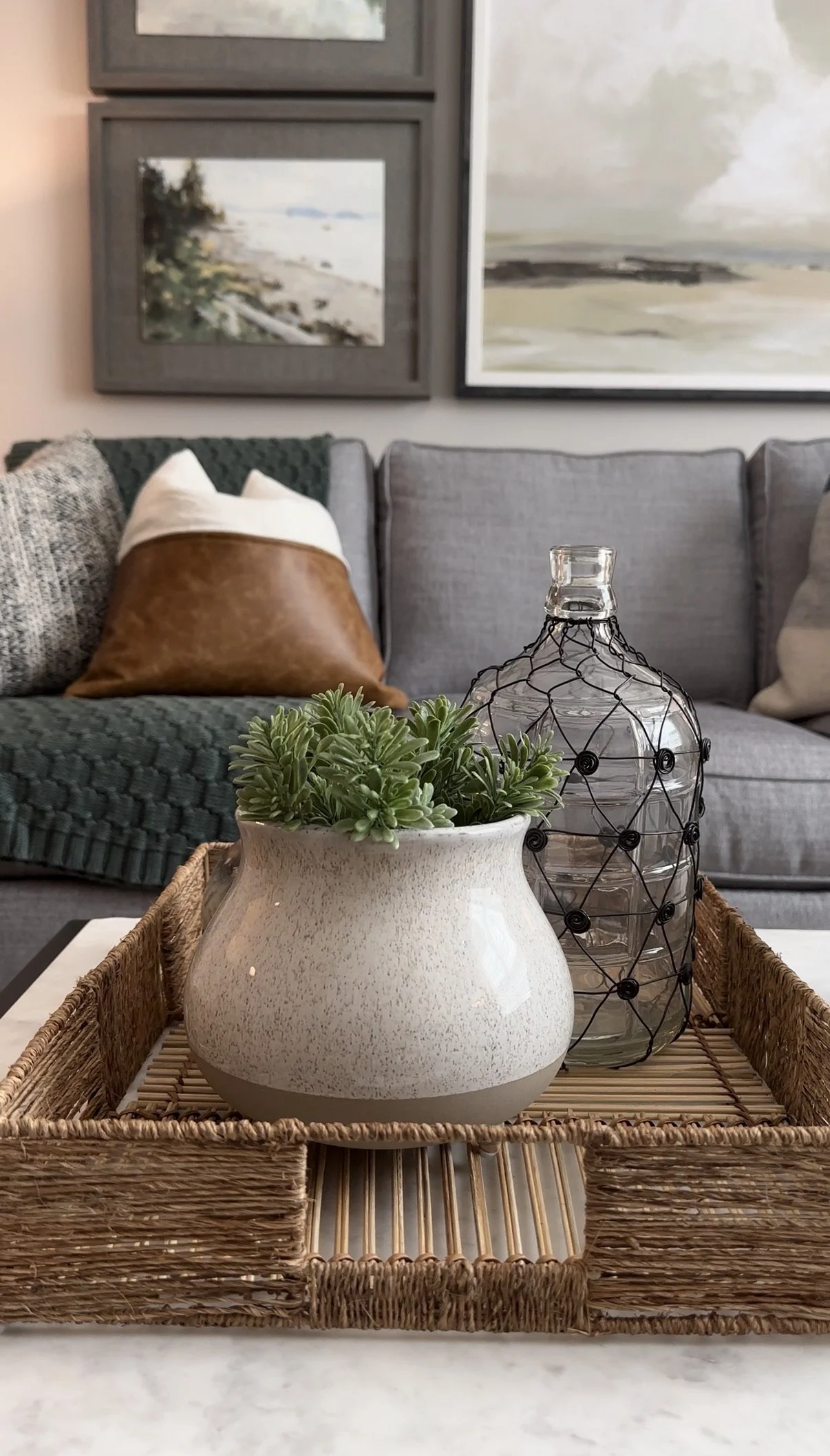 Close-up of decorative items on a tray on a table: Large white ceramic vase with green plant, and a glass bottle wrapped in black wire. In the background, a living room with a gray couch, pillows, and framed pictures on the wall.