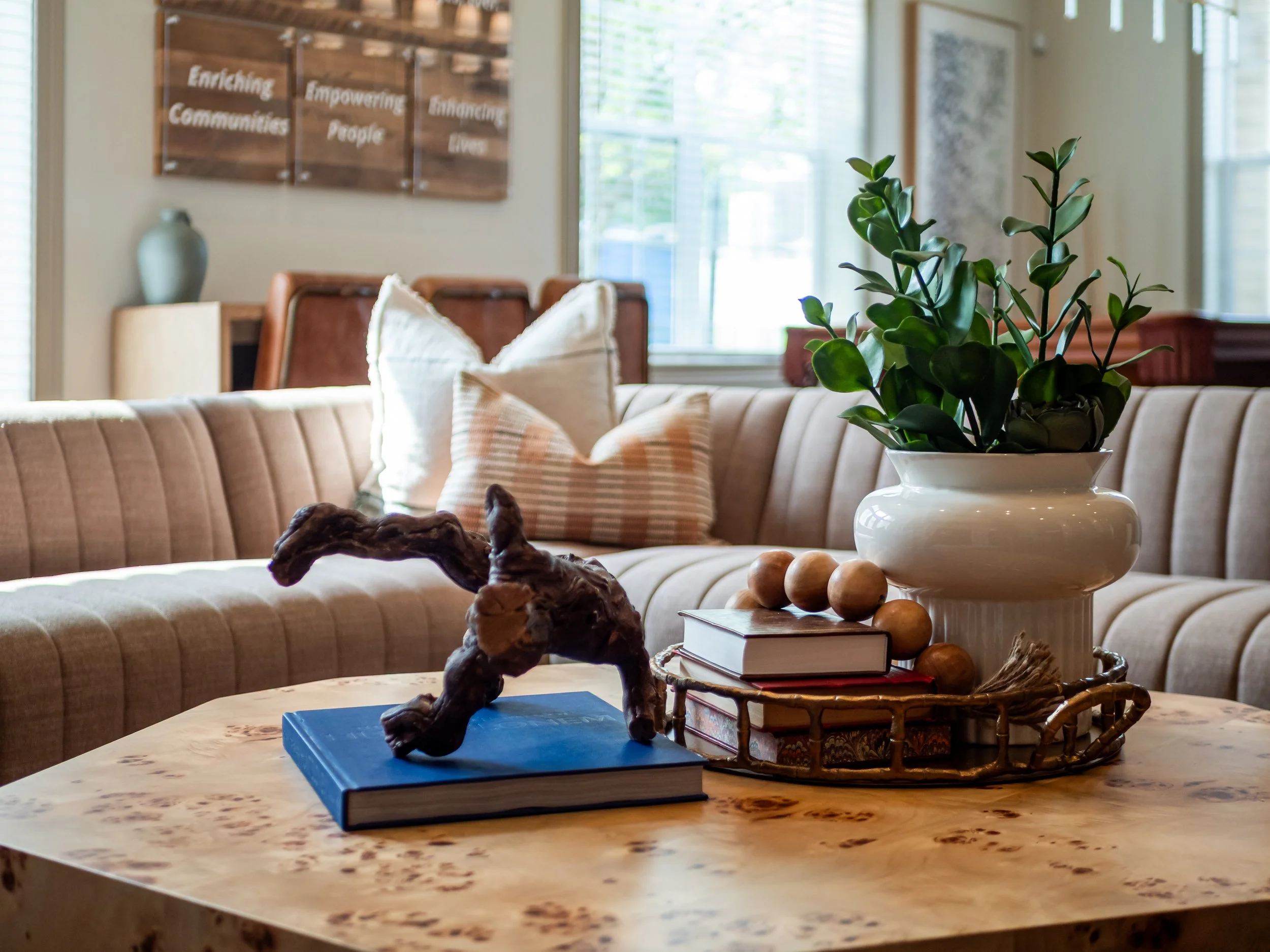 A living room with a beige sectional sofa adorned with pillows, a coffee table with decorative objects, a potted plant, and large windows allowing natural light. In the background, there is wall art with writing and a vase on a side table.