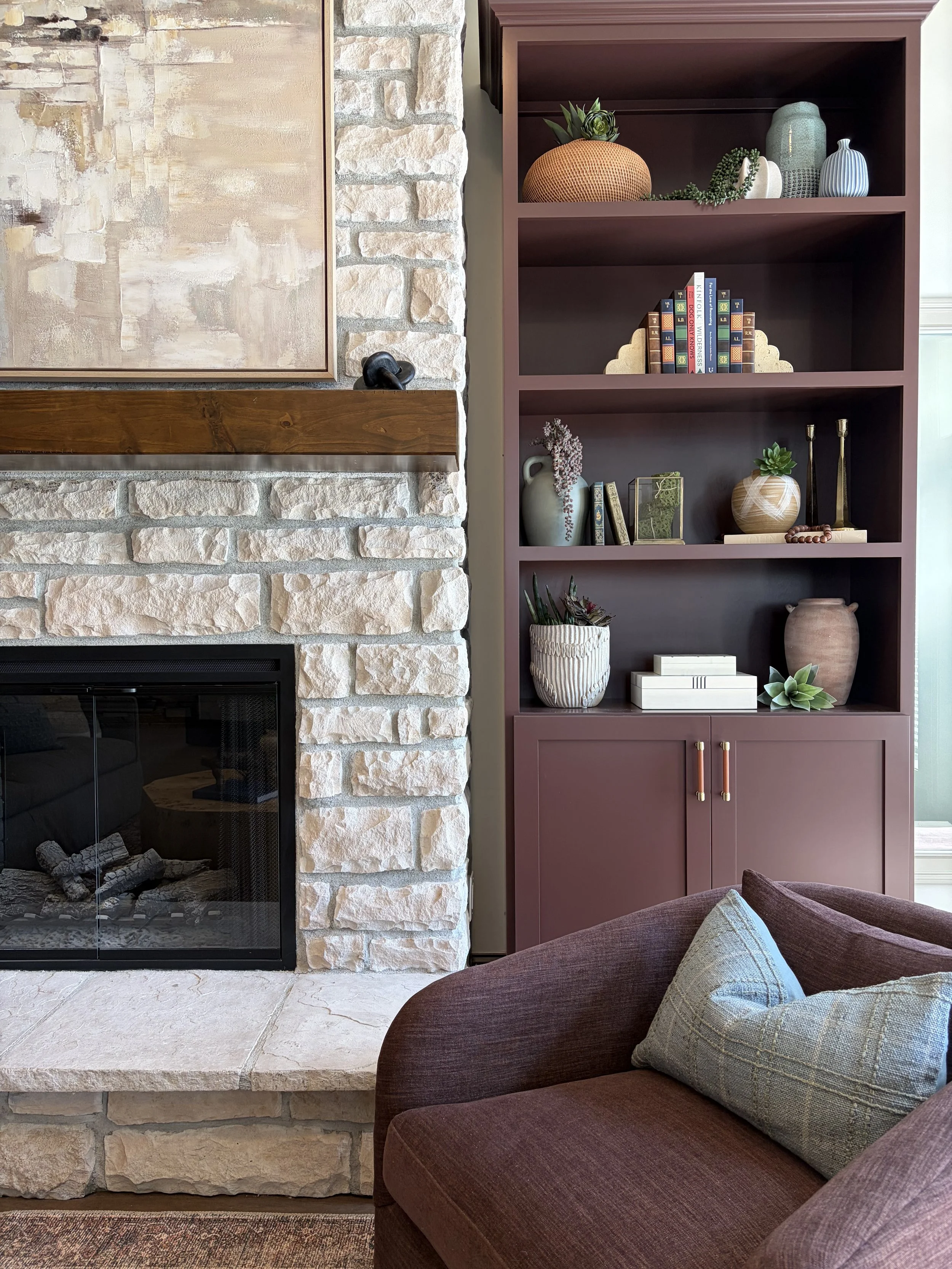 A living room corner featuring a brick and stone fireplace with a wooden mantel, a maroon bookshelf with decorative vases, books, and plants, and a maroon upholstered armchair with a patterned cushion.