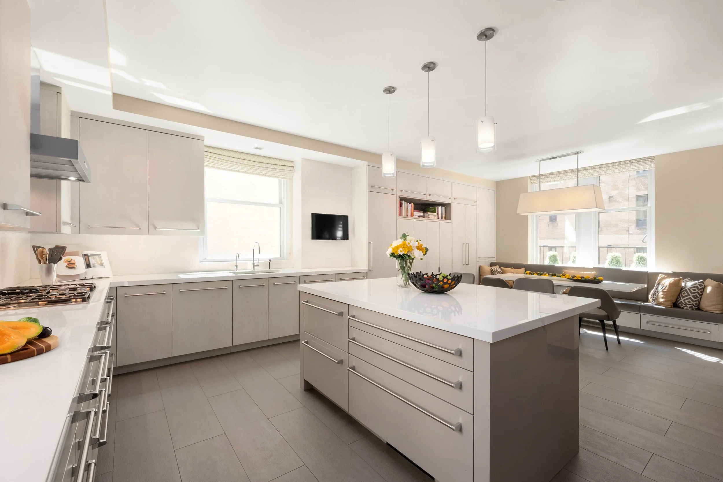 Modern kitchen with white cabinets, a white island, stainless steel appliances, a black wall-mounted TV, and large windows letting in natural light.