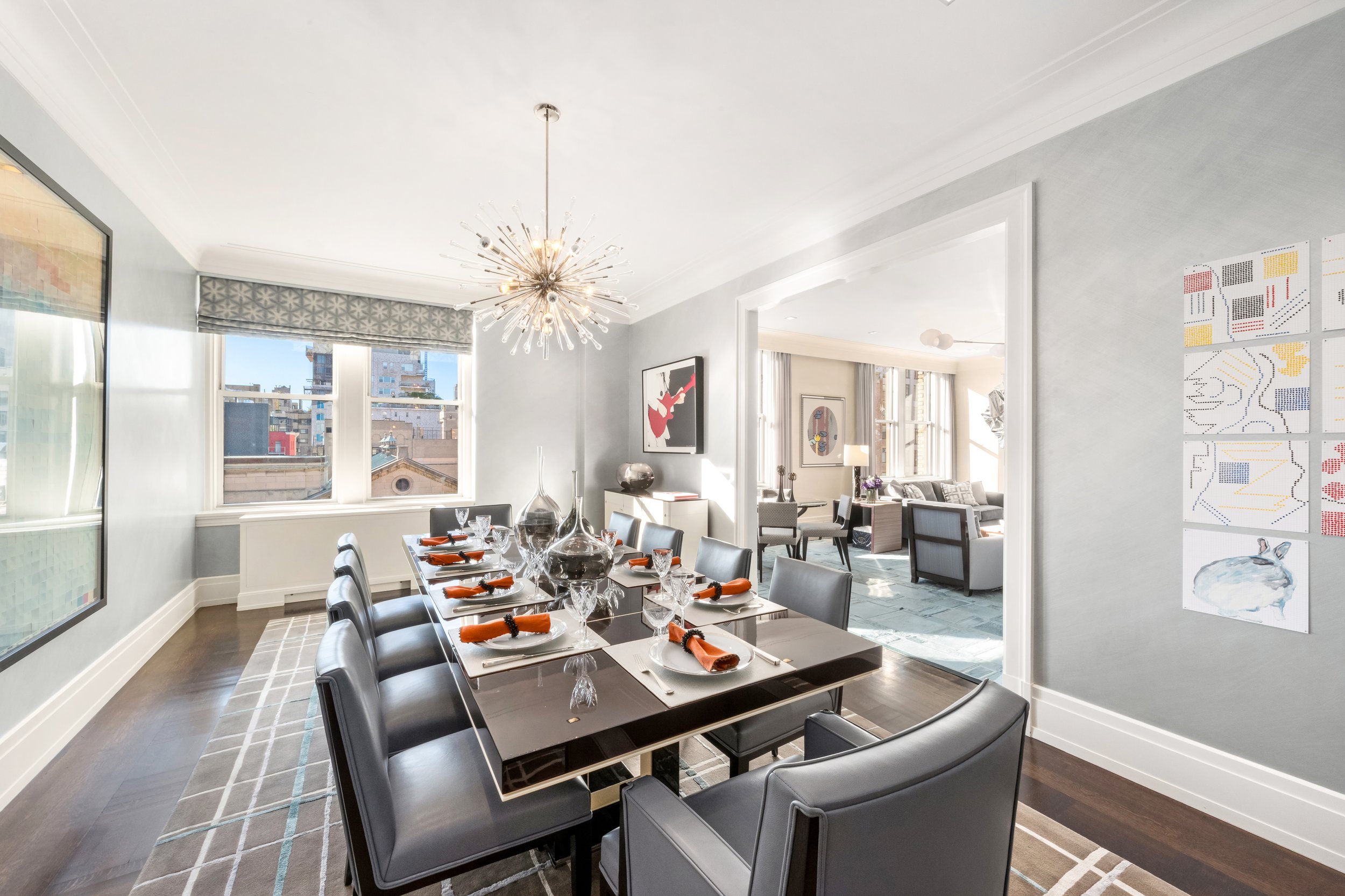A modern dining room with a large table set with orange napkins, wine glasses, and decorative vases, adjacent to a living area with artwork and natural light from windows.
