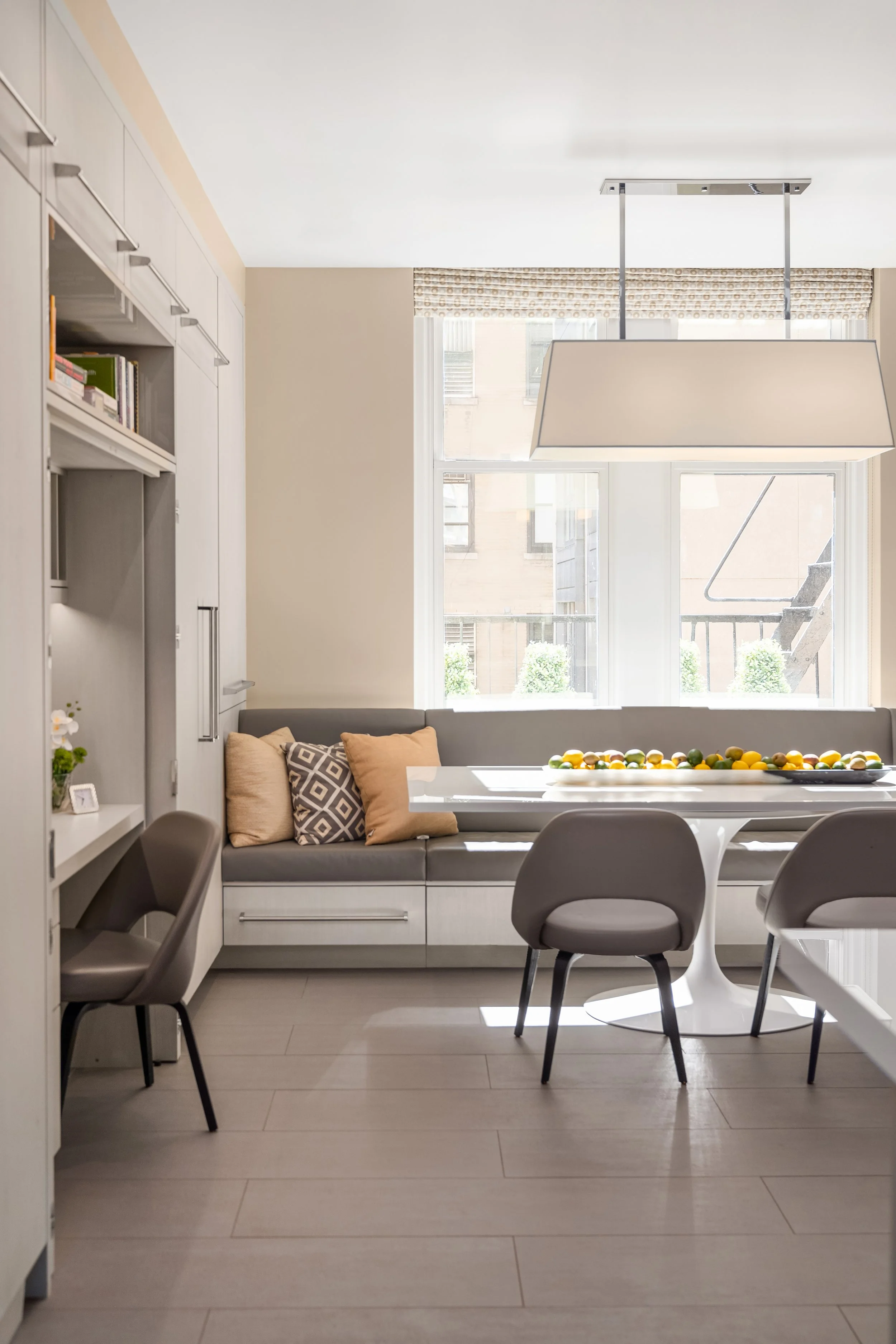 Modern dining room with a white table surrounded by four gray chairs, a built-in white sofa with pillows, and a window with a beige patterned valance, natural light illuminating the space.