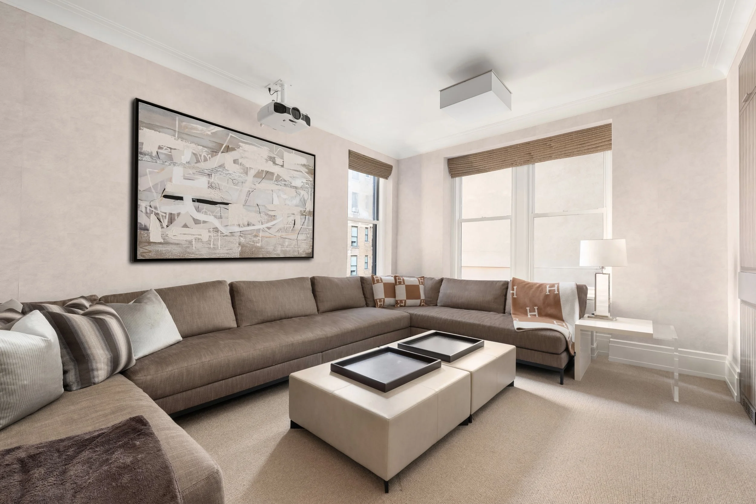 A living room with a large beige sectional sofa, multiple decorative pillows, and a white coffee table with three black trays. The room has light-colored walls, large windows with beige shades, a white ceiling with a ceiling-mounted projector, a mode