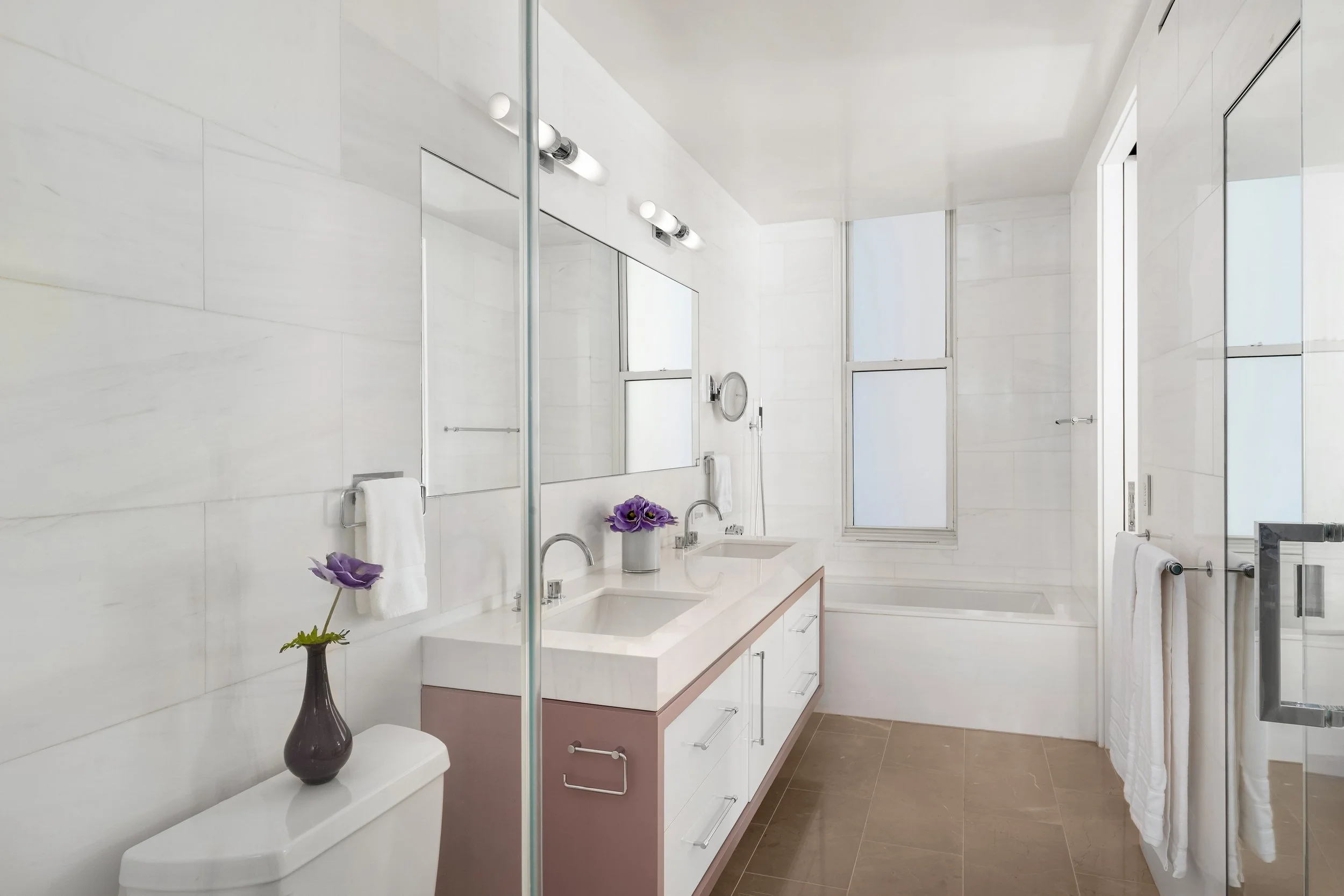 A modern, minimalist bathroom with white marble walls, a double sink vanity with a pink base, and large windows. There are purple flowers in a vase on the vanity and a large mirror above it. The bathroom includes a walk-in shower with a glass door, a