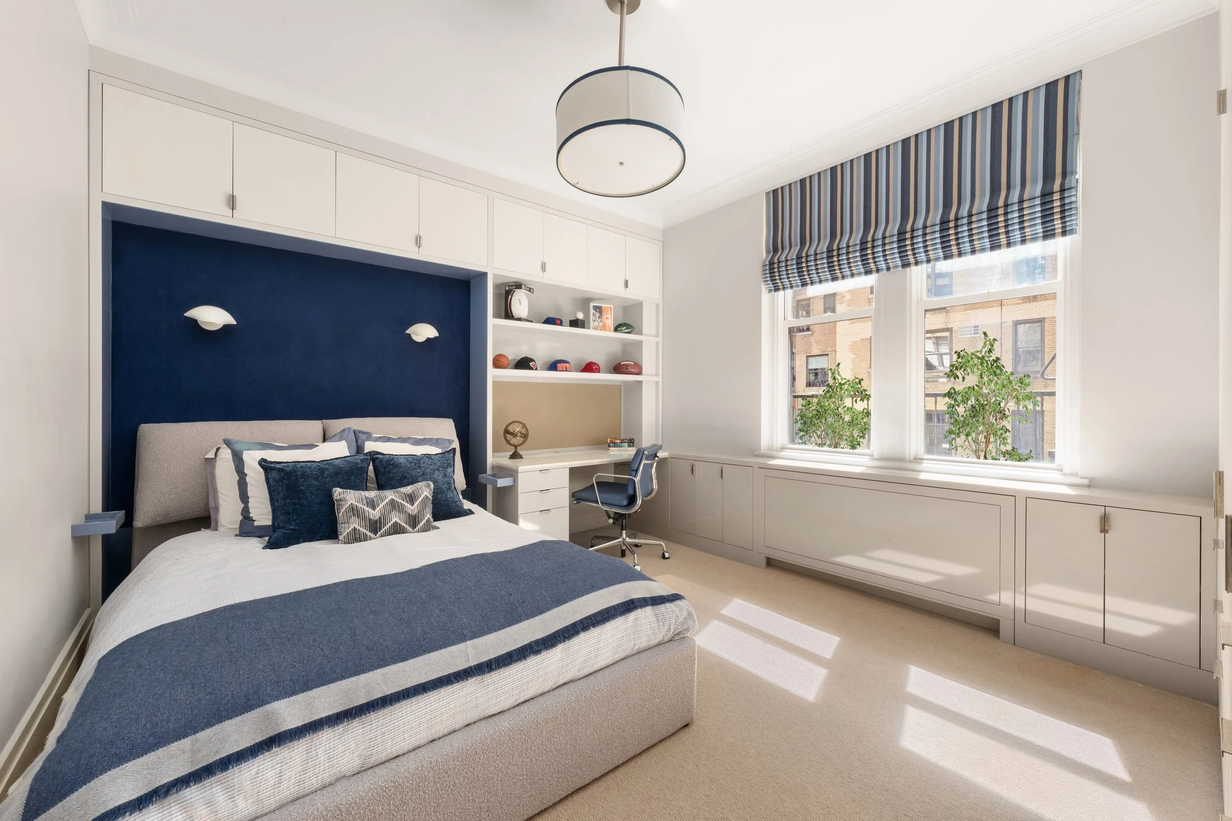 A cozy bedroom with a large bed that has a striped blue and white bedspread and numerous pillows. There is a navy wall behind the bed with two small white wall sconces. To the right, there is a built-in white desk with shelves above it, holding sport