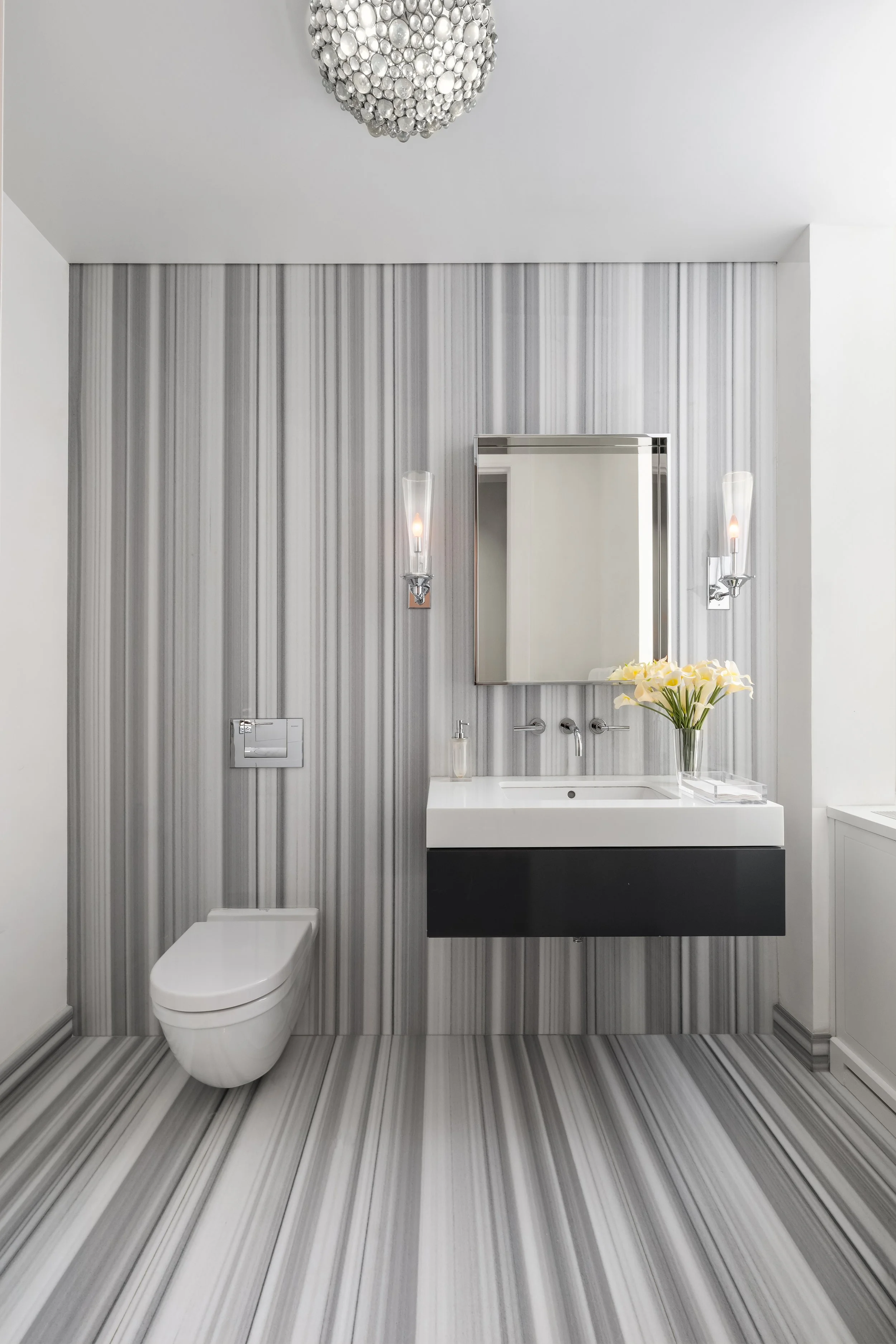 Modern bathroom with striped gray and white floor and wall tiles, a wall-mounted toilet, a white sink with a black cabinet, a rectangular mirror, and a vase with white flowers. There are wall-mounted light fixtures on either side of the mirror and a decorative spherical chandelier on the ceiling.