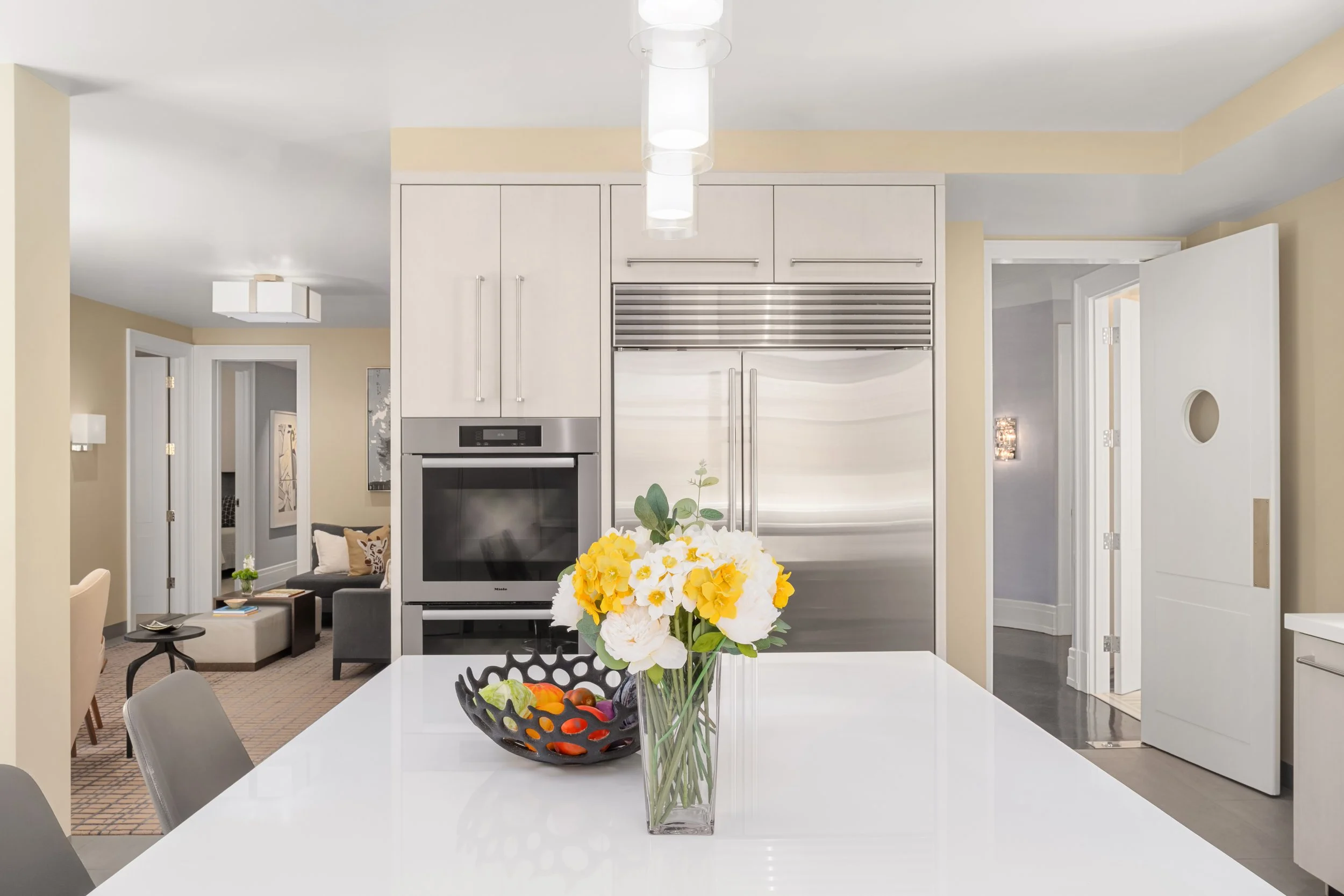 Modern kitchen with white dining table, yellow flowers in a glass vase, and a bowl of oranges and apples, with stainless steel refrigerator and built-in oven in the background.
