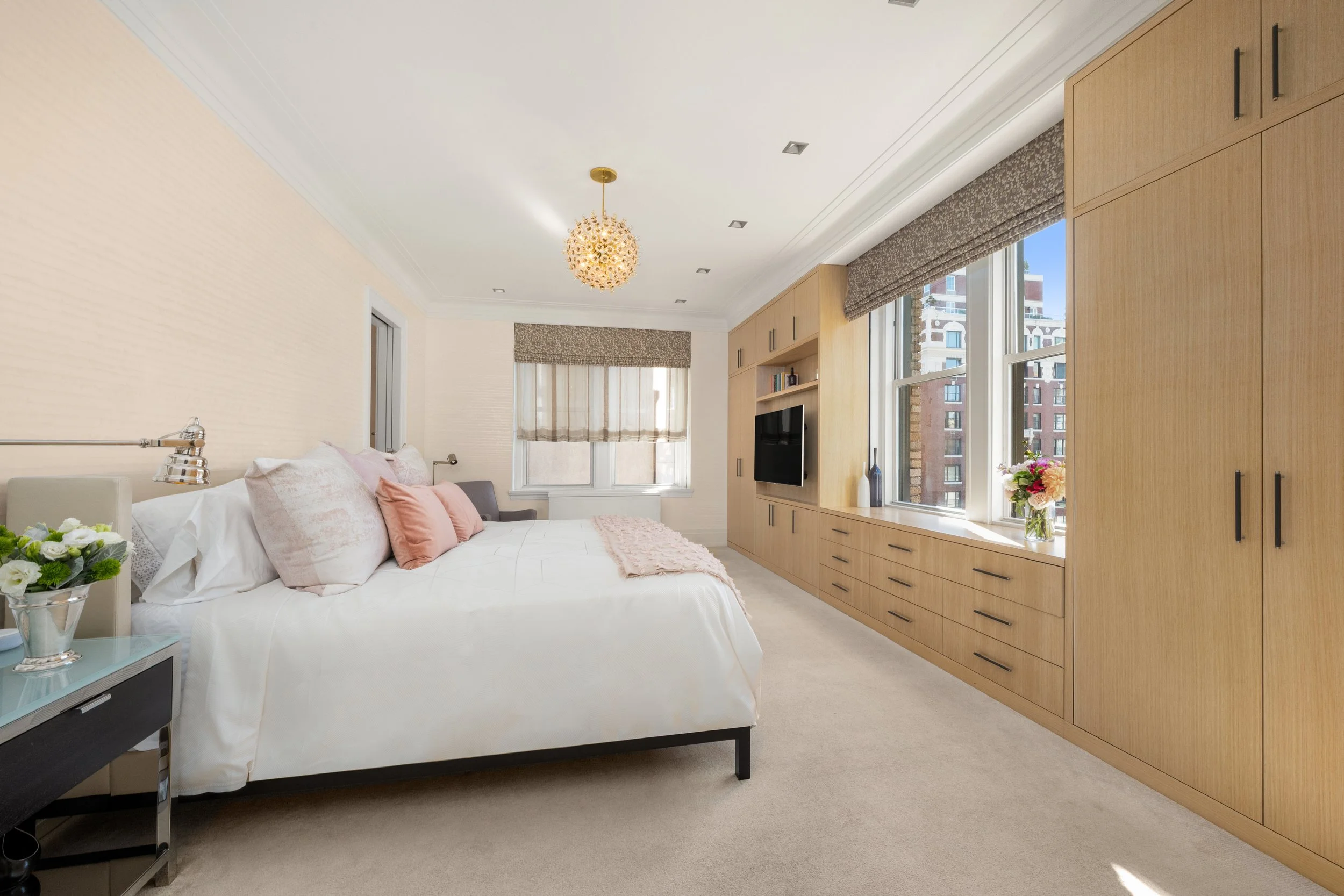A bedroom with a large bed, wooden built-in cabinets, a window with beige shades, and a modern chandelier hanging from the ceiling.