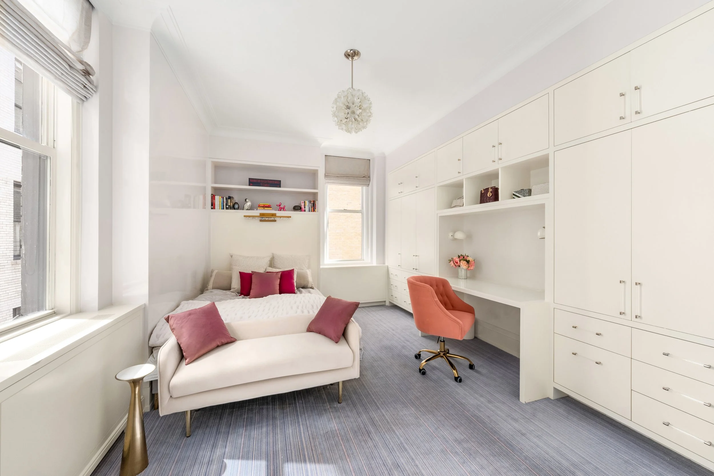 Bright bedroom with white walls and built-in white cabinets, a white upholstered bed with pink and burgundy pillows, a pink swivel chair, a window with blinds, and a floral chandelier.