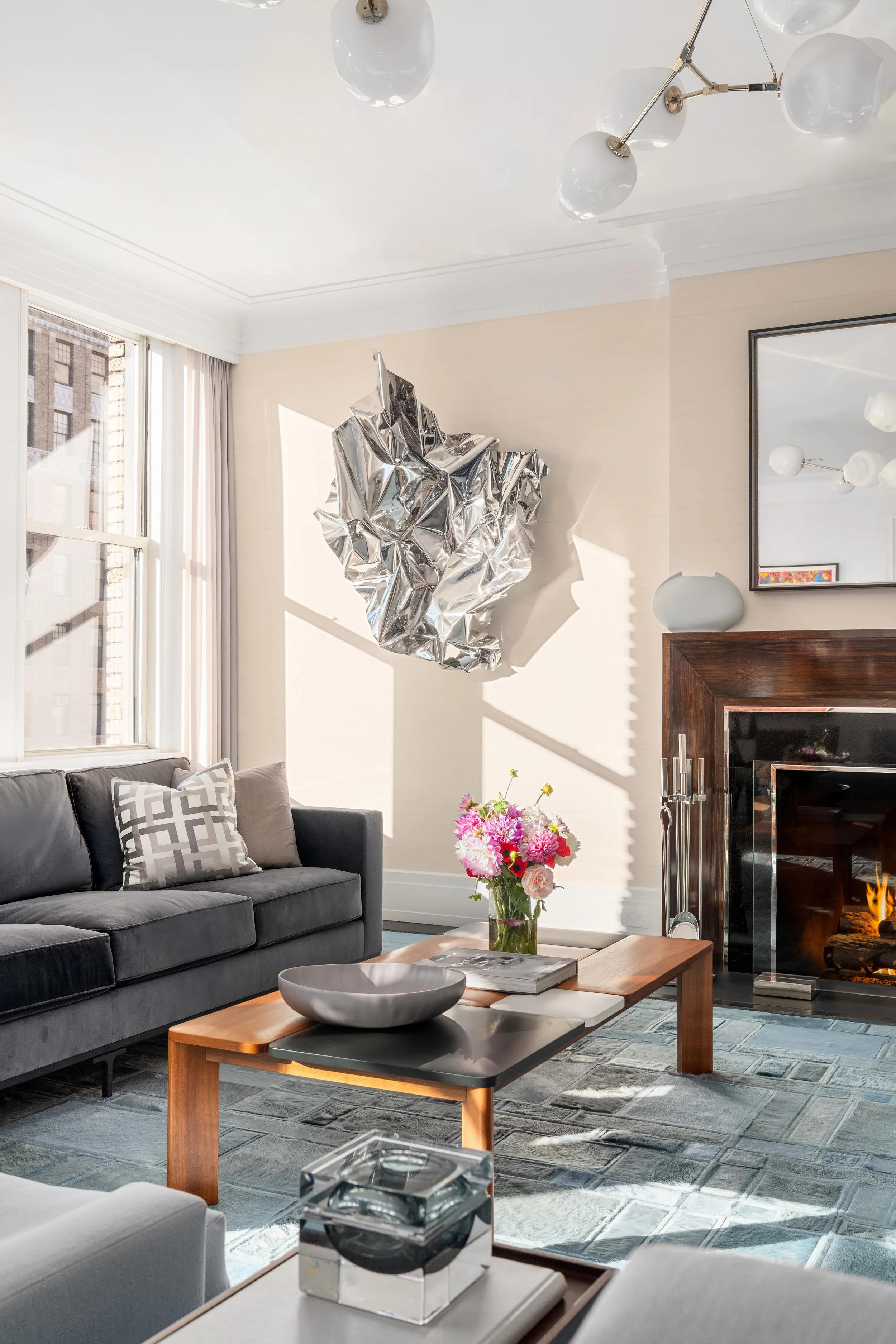 Bright living room with a gray sofa, a wooden coffee table with a floral arrangement, a large mirror, and a modern chandelier. Sunlight streams through large windows, casting shadows on the wall.
