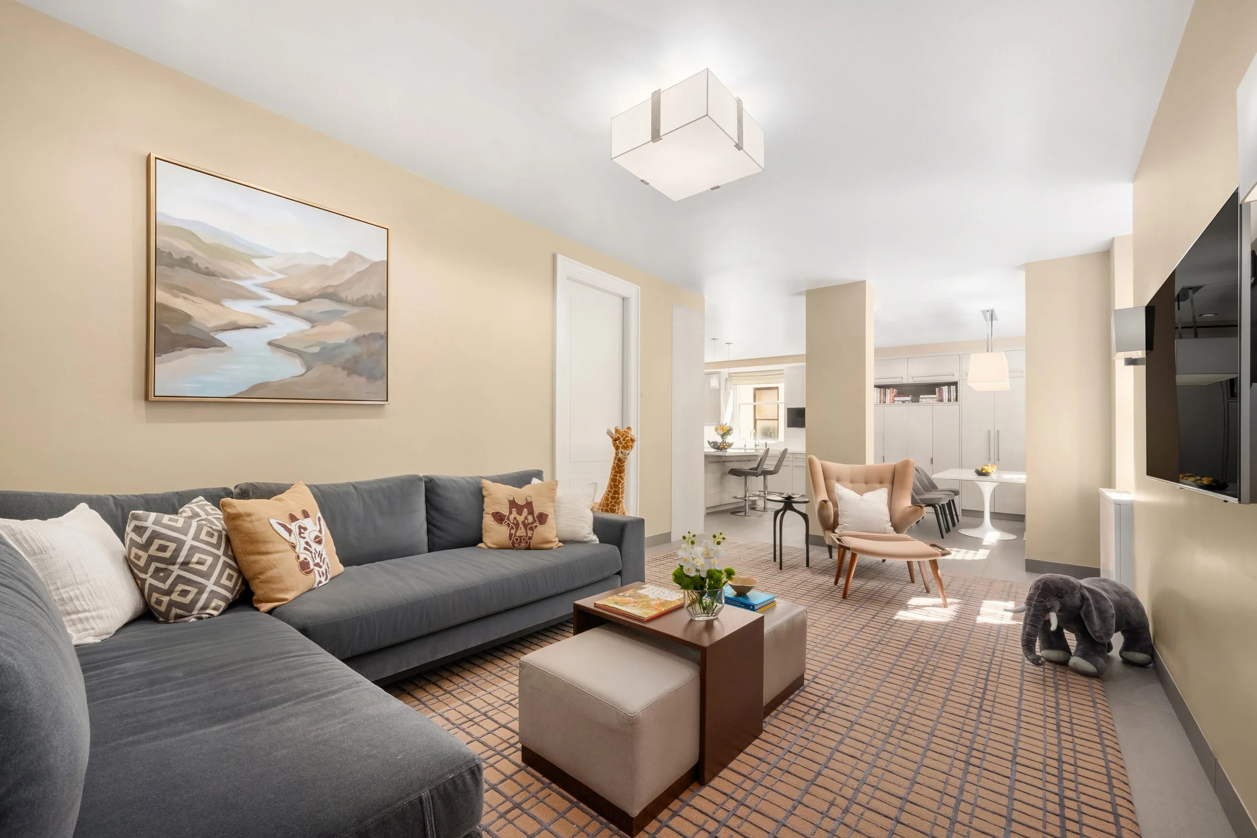 A spacious living room with a large gray sofa decorated with animal print pillows, a small wooden coffee table, an upholstered beige armchair, a baby elephant plush toy, artwork of a river and mountains on the wall, and a view into the bright kitchen