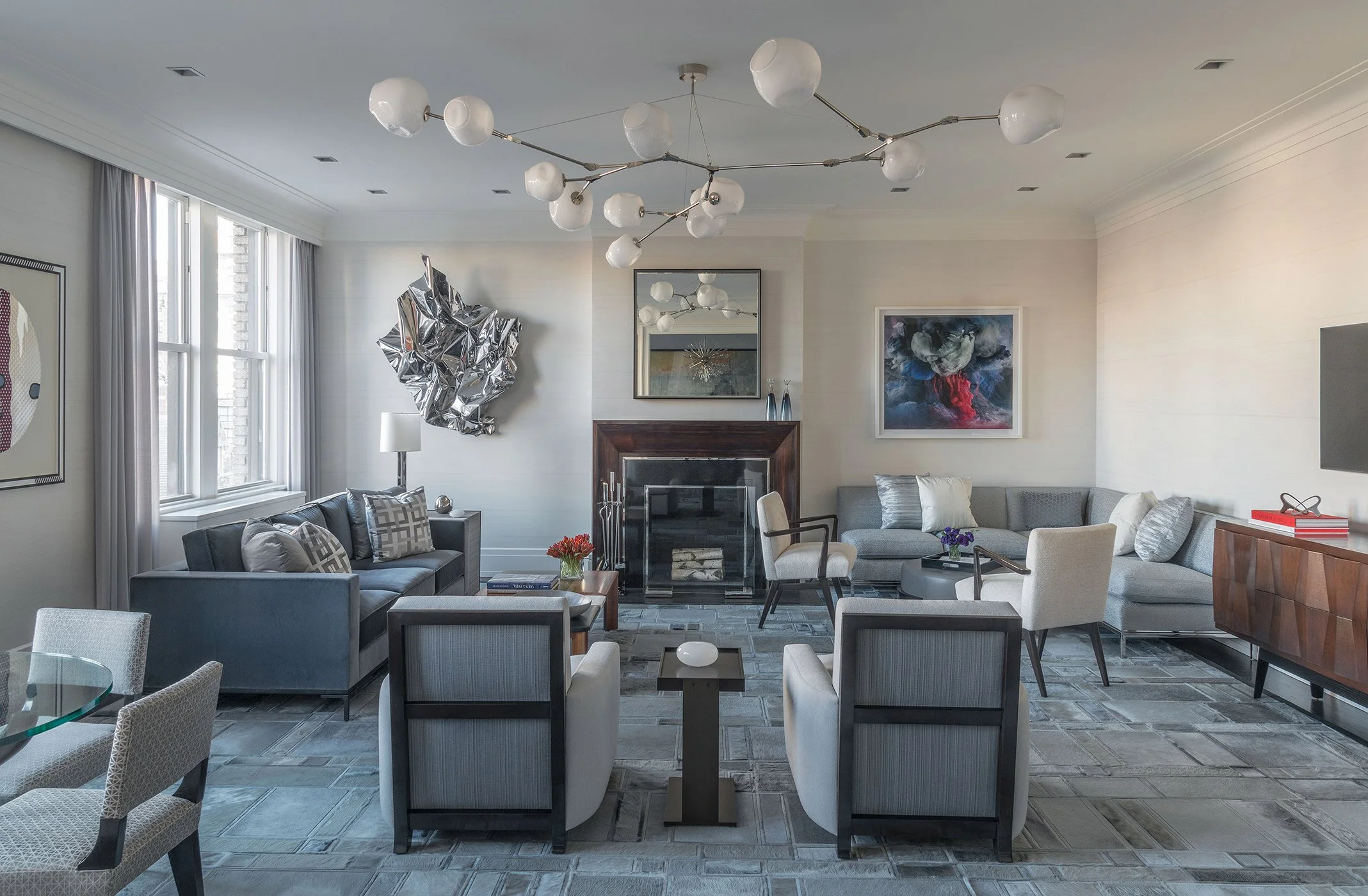 Modern living room with white walls, a gray sofa, and white armchairs. There are decorative pillows, a glass-topped coffee table, and artwork on the walls. A ceiling fixture with multiple white globes hangs above, with windows covered by white curtai
