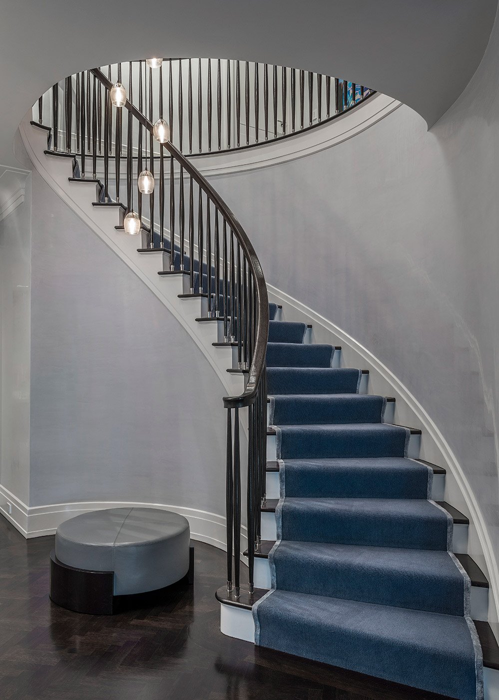 Interior of a home featuring a curved staircase with a dark wooden handrail, blue carpeted steps, black balusters, modern hanging light fixtures, gray walls, a dark wood floor, and a round gray ottoman.
