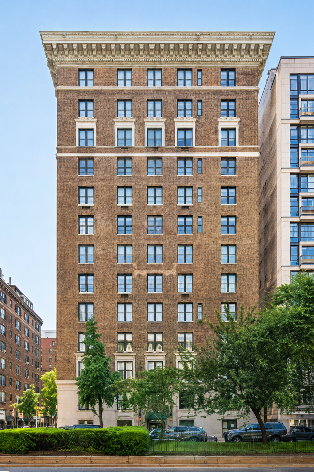 A tall brick residential building with decorative white trim and numerous windows, surrounded by trees and parked cars, under a clear blue sky.