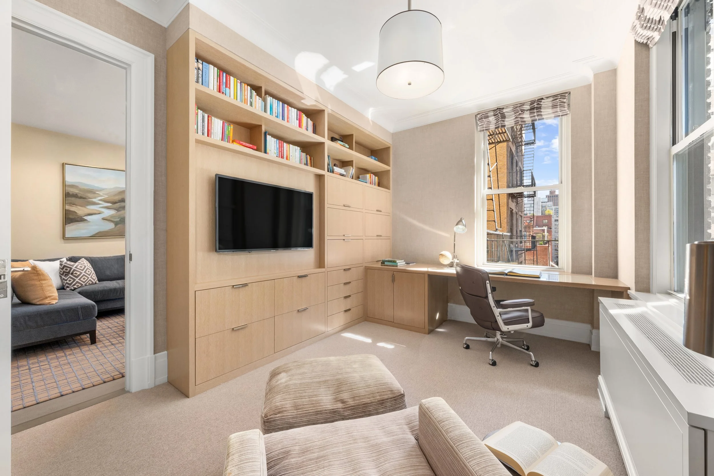 A home office with a desk, a swivel chair, and a built-in wooden bookshelf with a TV. There's a large window with city buildings outside, and sunlight streaming in. A beige armchair and an open book are also visible.