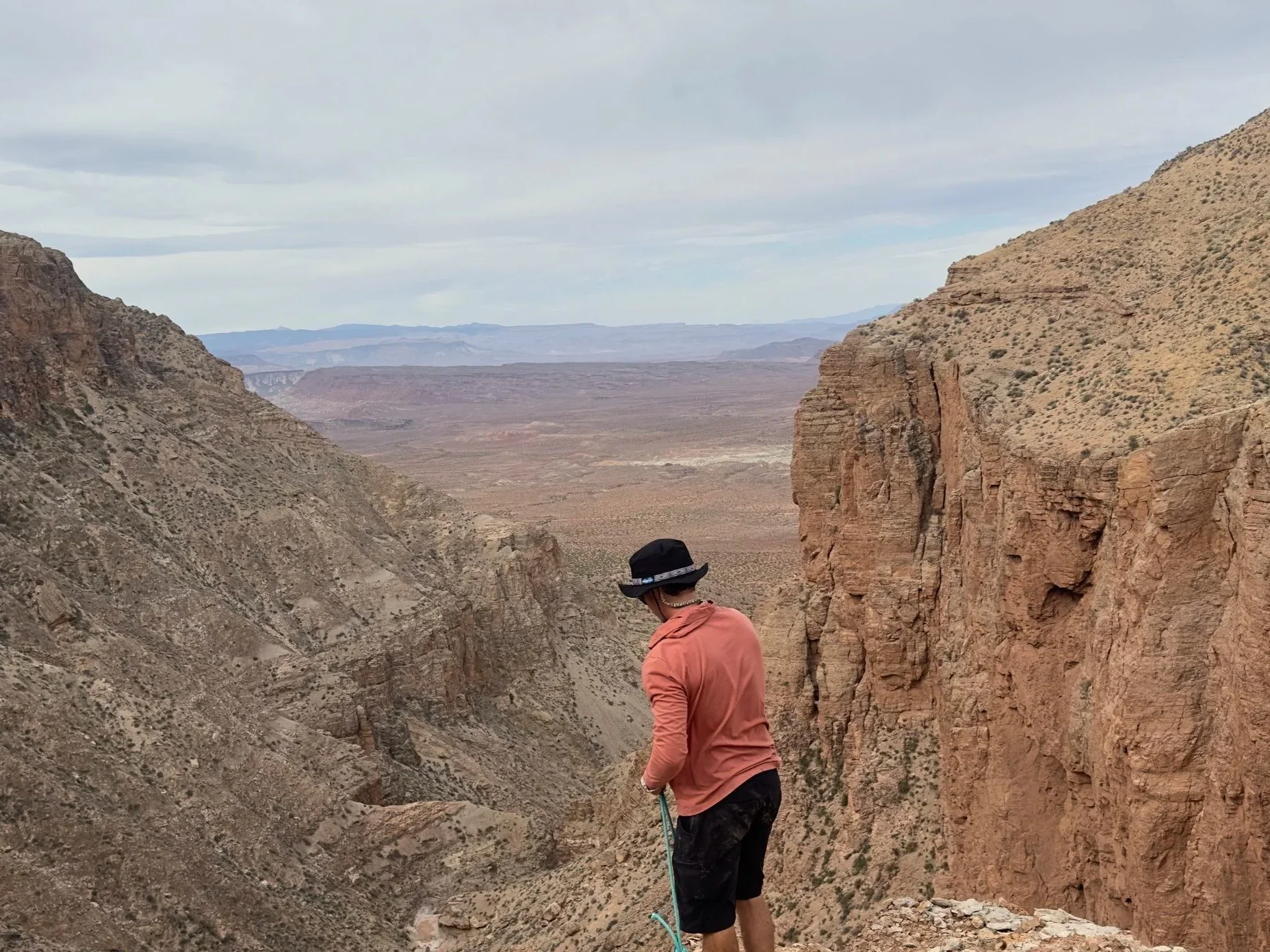 A man wearing a black hat, orange jacket, and black shorts stands on a rocky canyon ledge holding a green rope, with a vast desert landscape and canyon walls in the background under a cloudy sky.