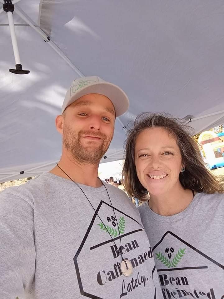 A smiling man and woman taking a selfie at an outdoor event under a white canopy tent, both wearing gray T-shirts with "Bean Caffeinated Lately" and a coffee bean logo.